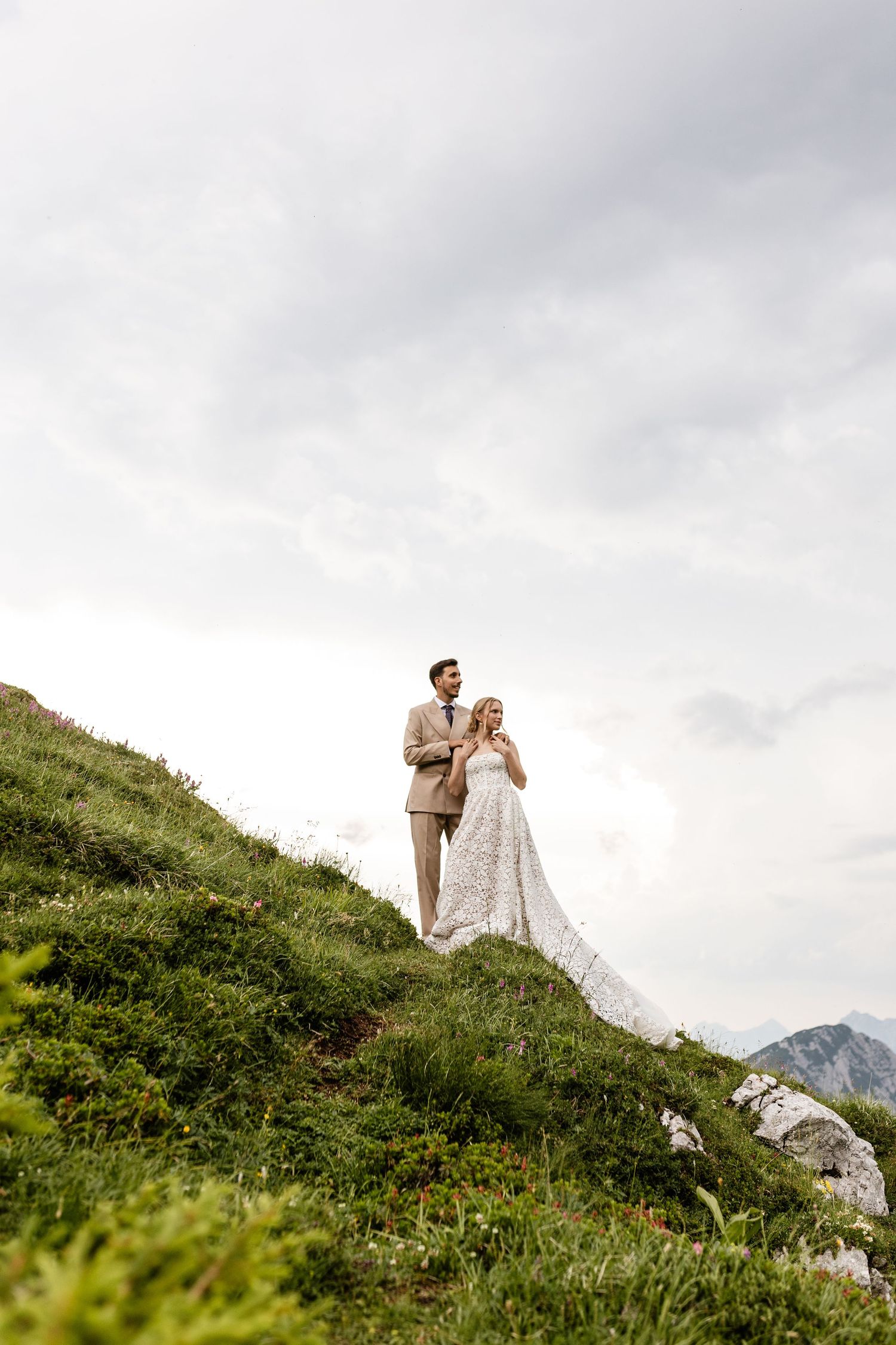 Bride in lace gown and groom in tan suit stand together on lush green Alpine hillside under cloudy sky.