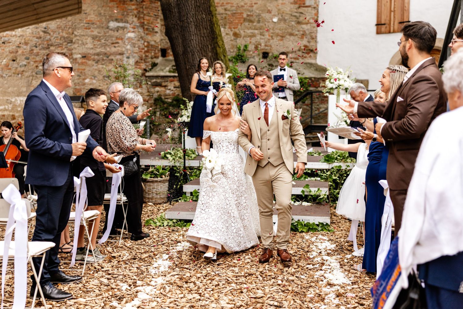 Bride and groom walk down outdoor aisle as guests toss petals at rustic wedding ceremony.