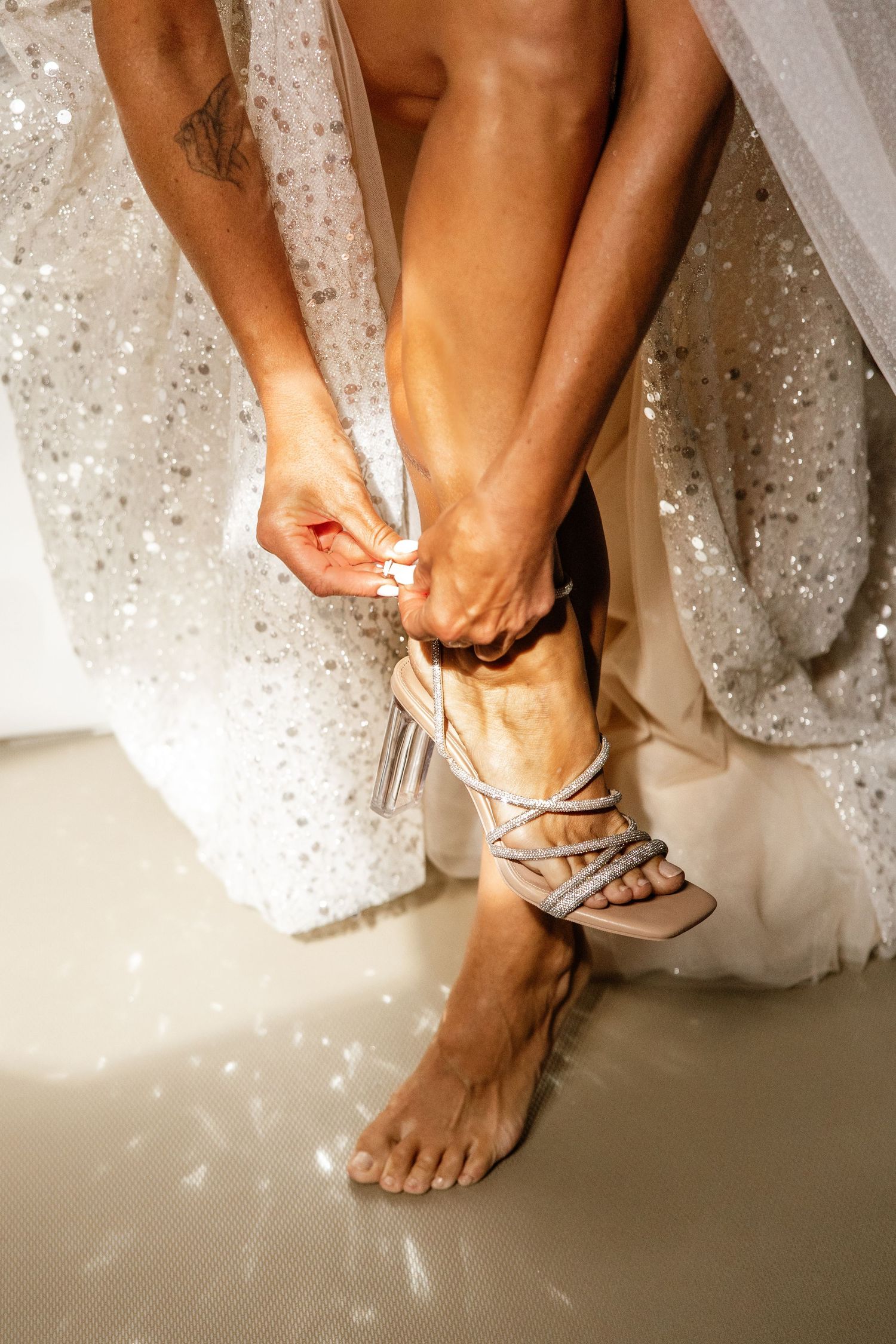 Bride fastening strappy silver heeled sandals while wearing a sparkly white wedding dress.