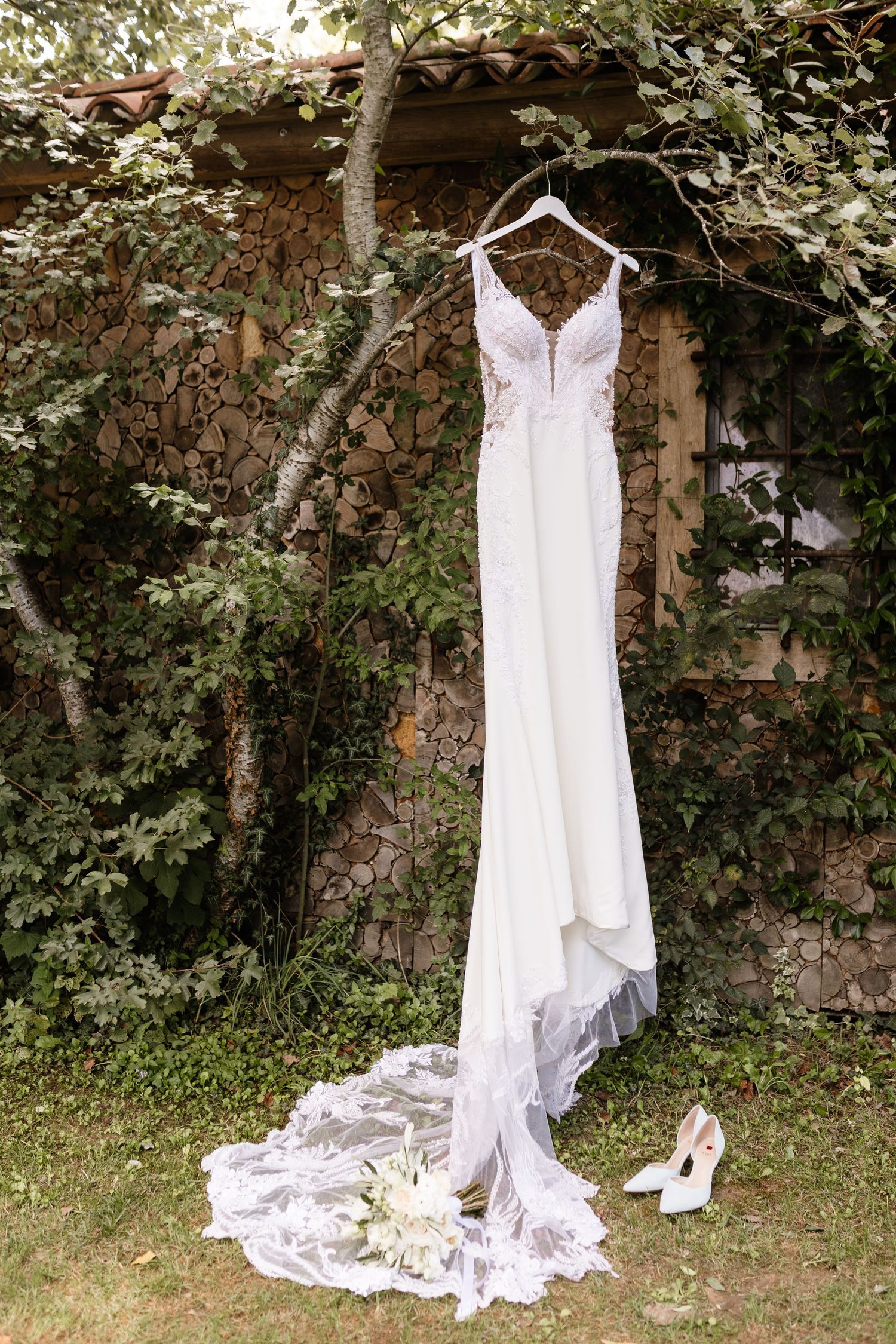 Elegant white wedding dress with lace train hanging on a tree branch outdoors with bridal heels and bouquet below.