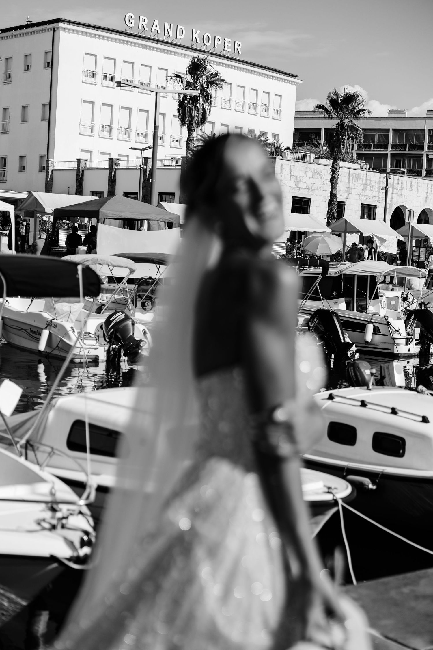 Black and white photo of a bride in a wedding dress near a marina with boats and Grand Koper hotel in background.