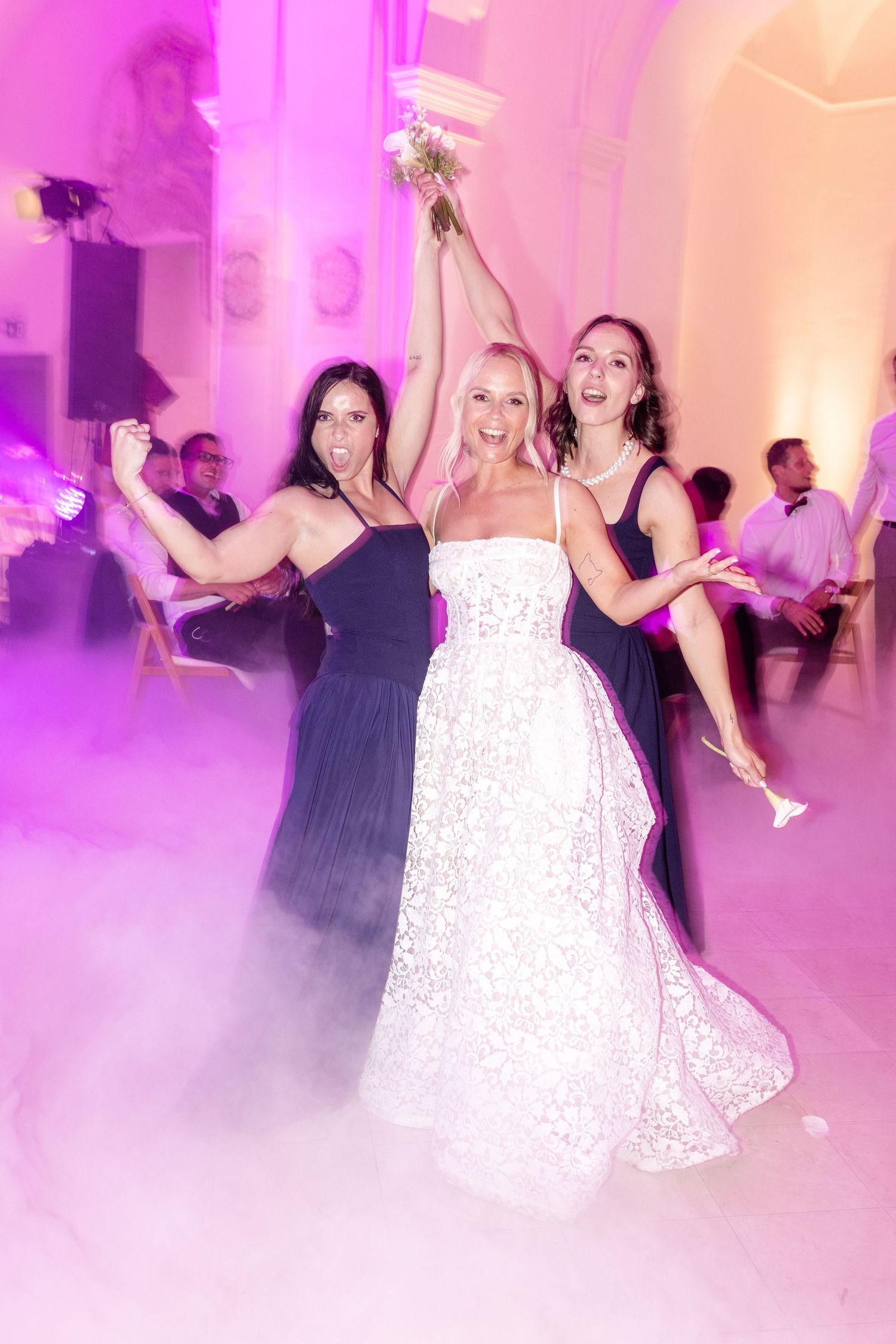 Joyful bride in white lace gown raising bouquet with two bridesmaids in navy dresses on foggy pink dance floor.