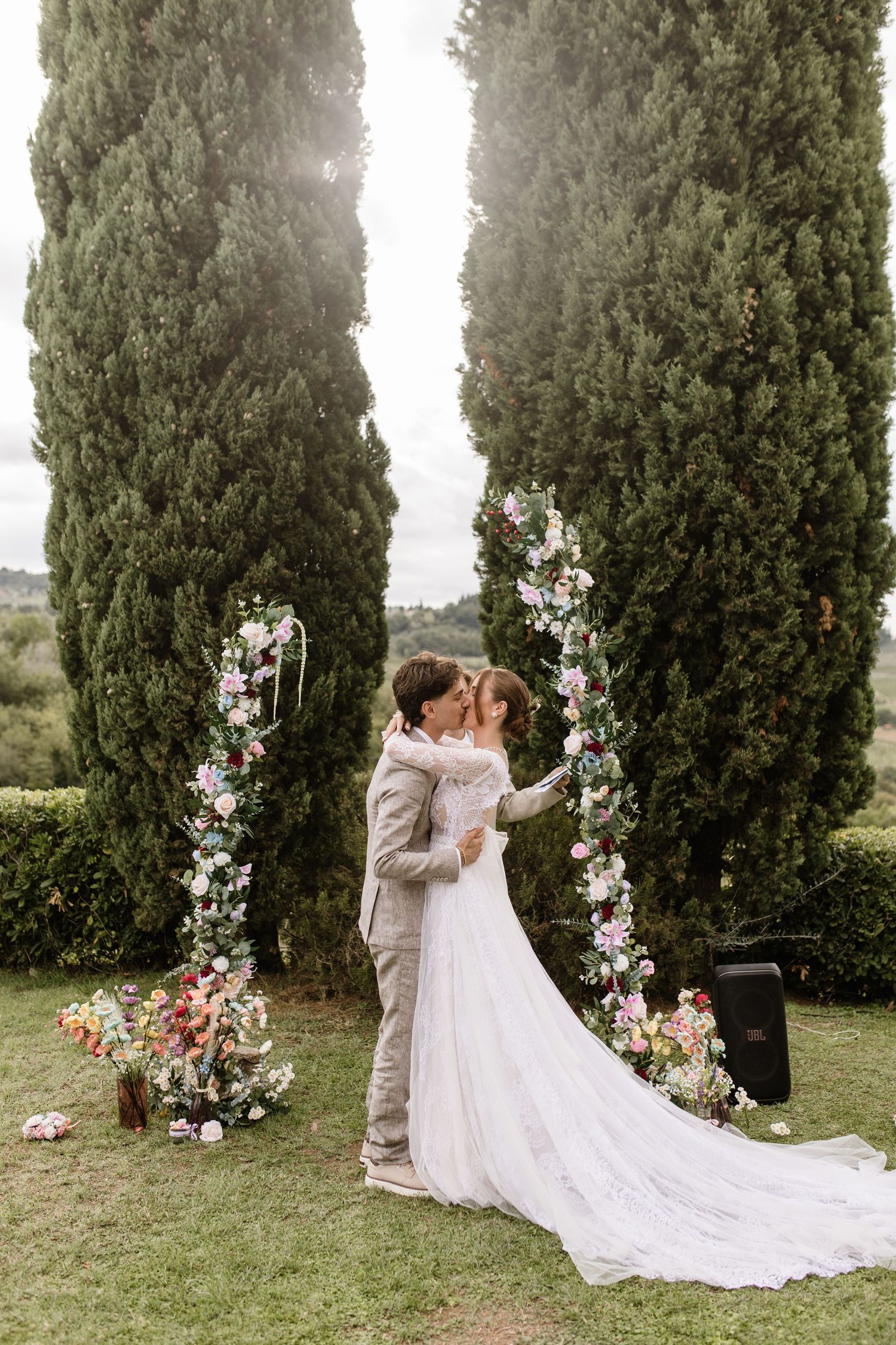 Bride and groom sharing a kiss under a floral arch with tall cypress trees at a Tuscan wedding.