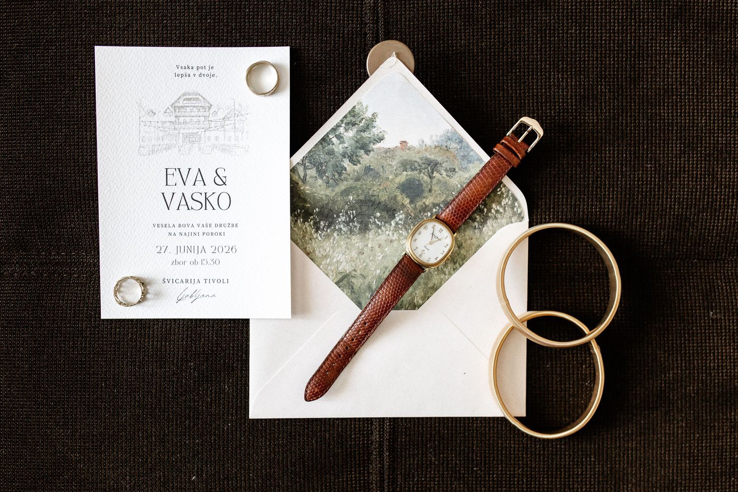 Wedding flat lay featuring Eva and Vasko invitation, brown leather watch, gold rings, and envelope with landscape photo.
