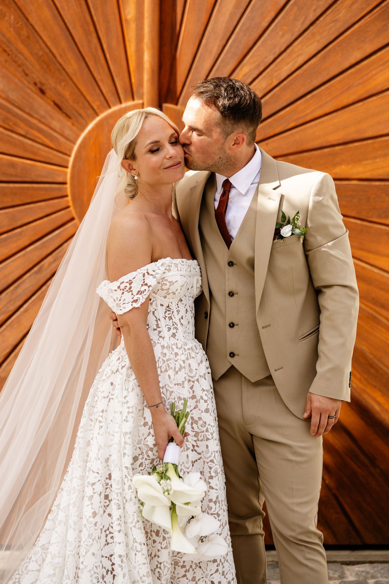 Groom in tan suit kissing bride in lace off-shoulder gown holding white calla lilies.
