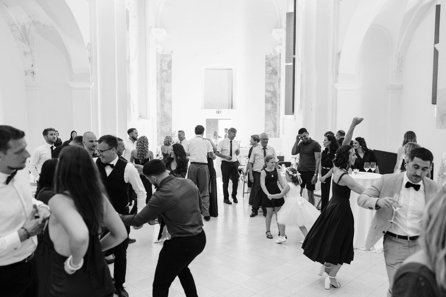 Guests dance joyfully at an elegant wedding reception in a grand white hall, black and white photography.