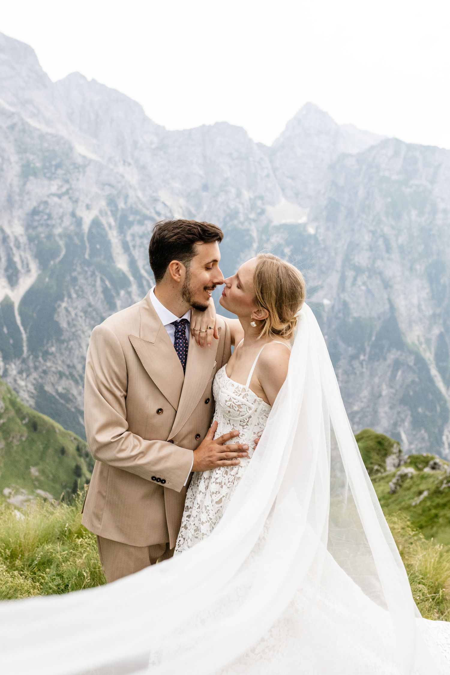 Bride and groom embrace before stunning mountain backdrop, her veil flowing in the wind on their wedding day.