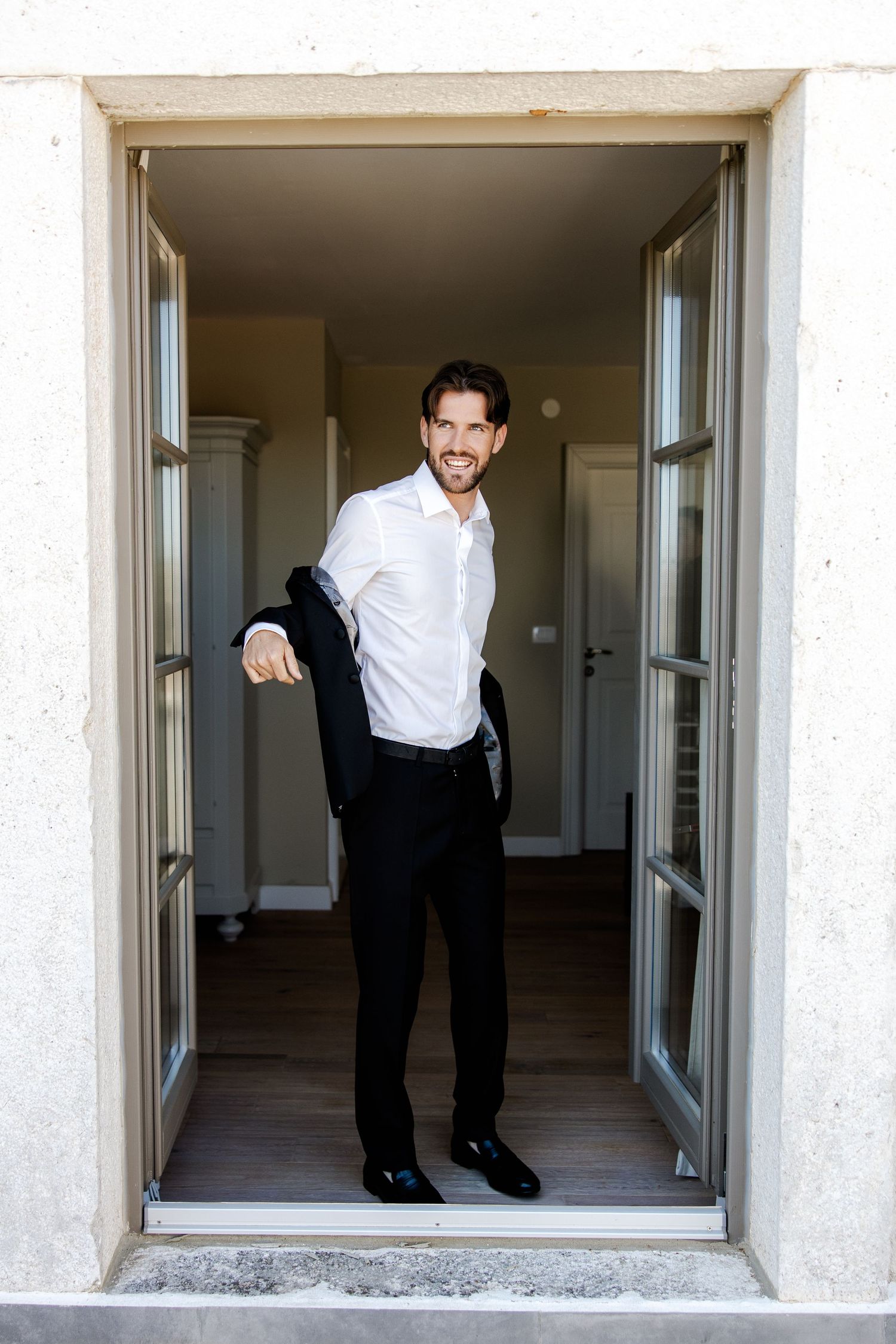 Smiling groom in white shirt holding black suit jacket standing in doorway.