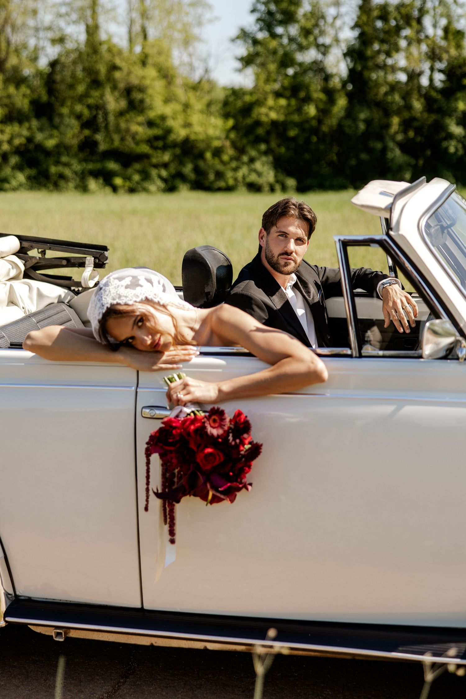 Bride and groom posing in a white vintage convertible car with a red rose bouquet on the door.