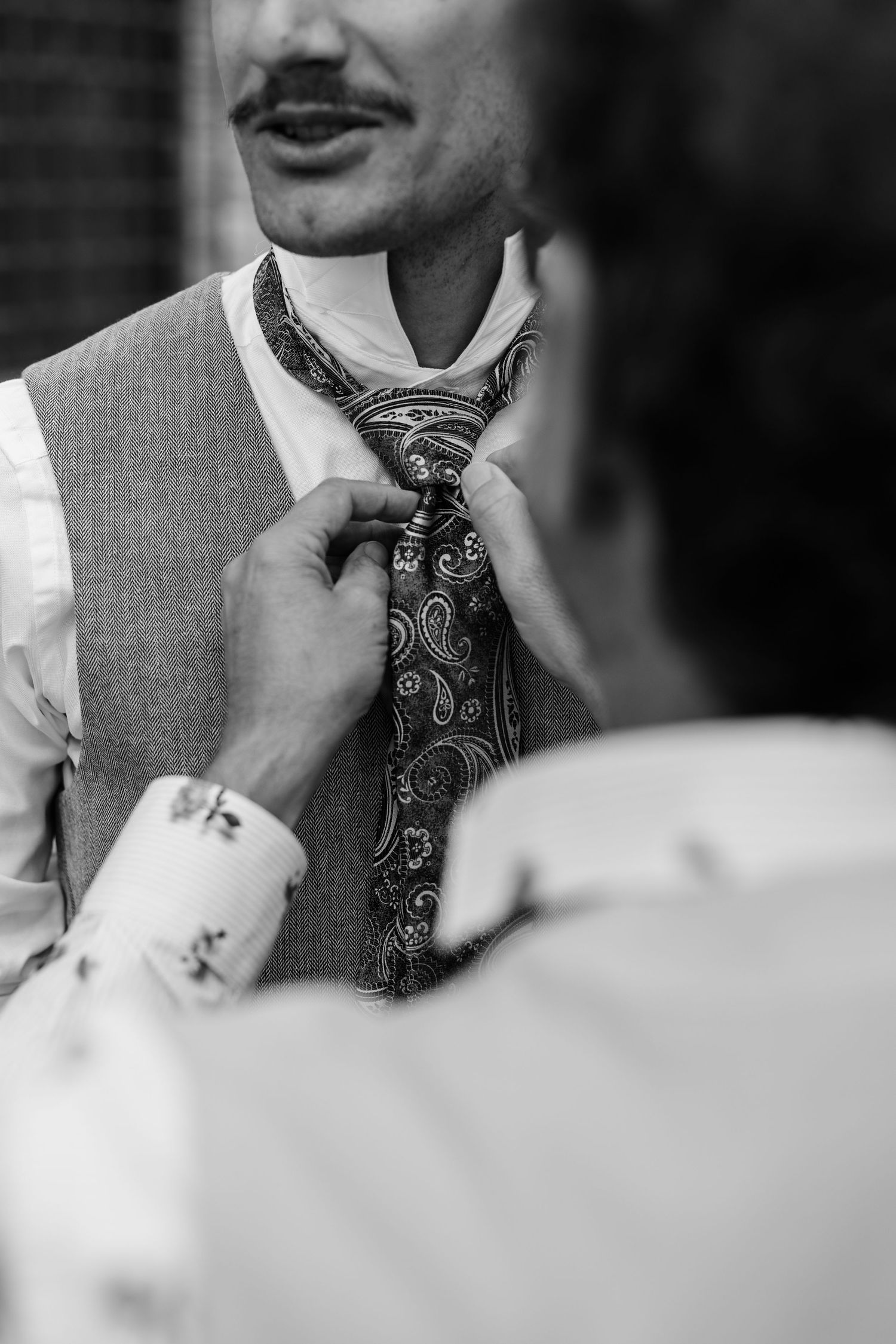 Groom with mustache adjusts paisley tie while someone helps him dress for wedding.