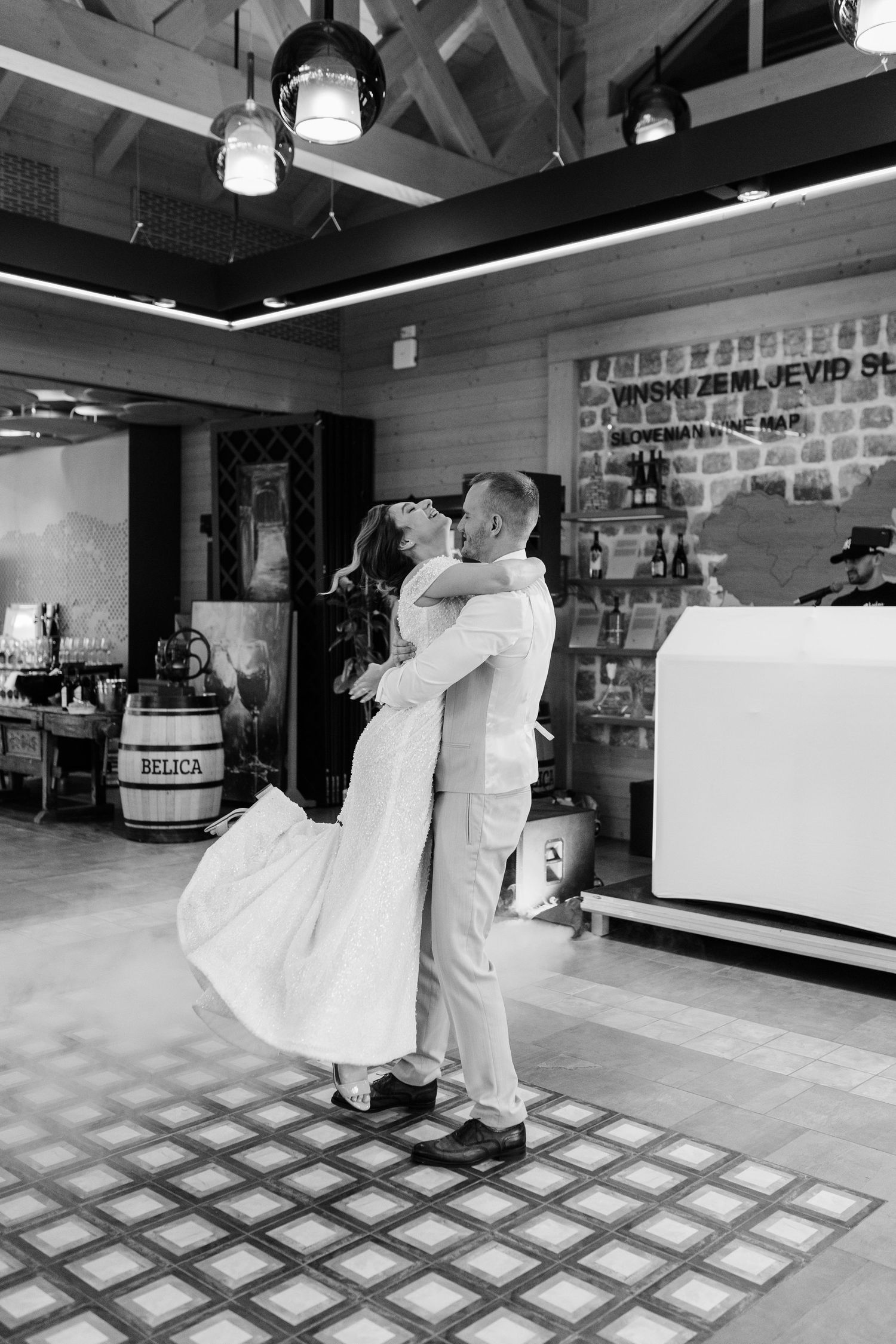 Groom lifts bride during first dance in a rustic venue, captured in elegant black and white photography.