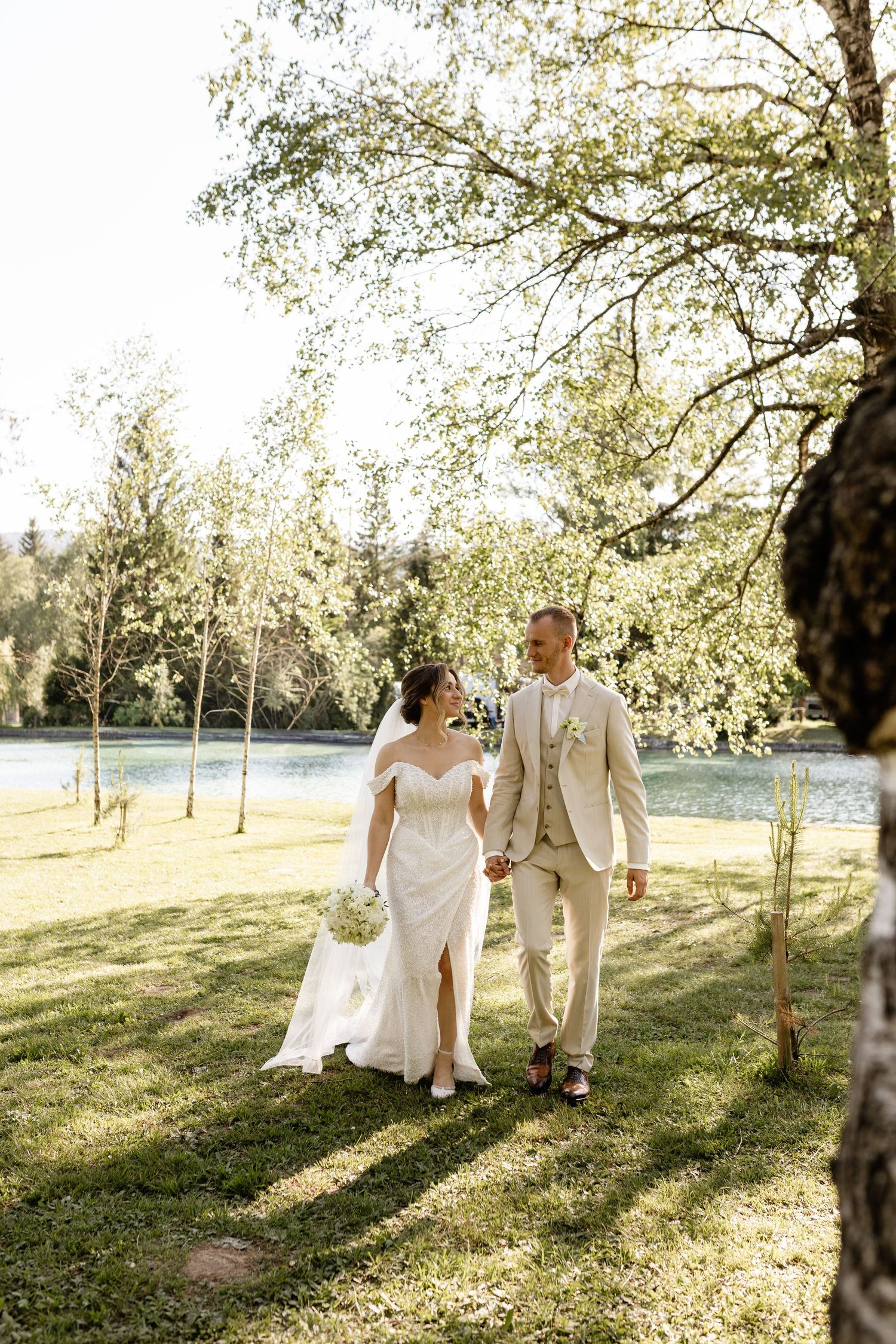 Bride and groom walk hand-in-hand through a sunlit garden near a lake, bride in white gown, groom in beige suit.