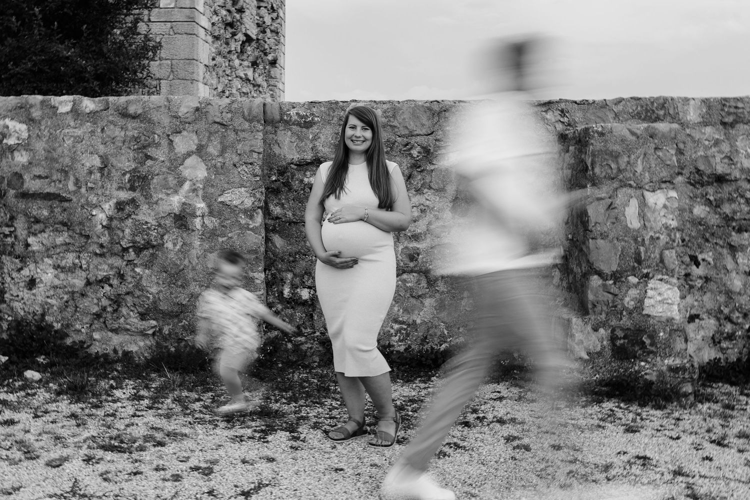 Black and white photo of a pregnant woman in a white dress posing outdoors near a stone wall with blurred figures nearby.