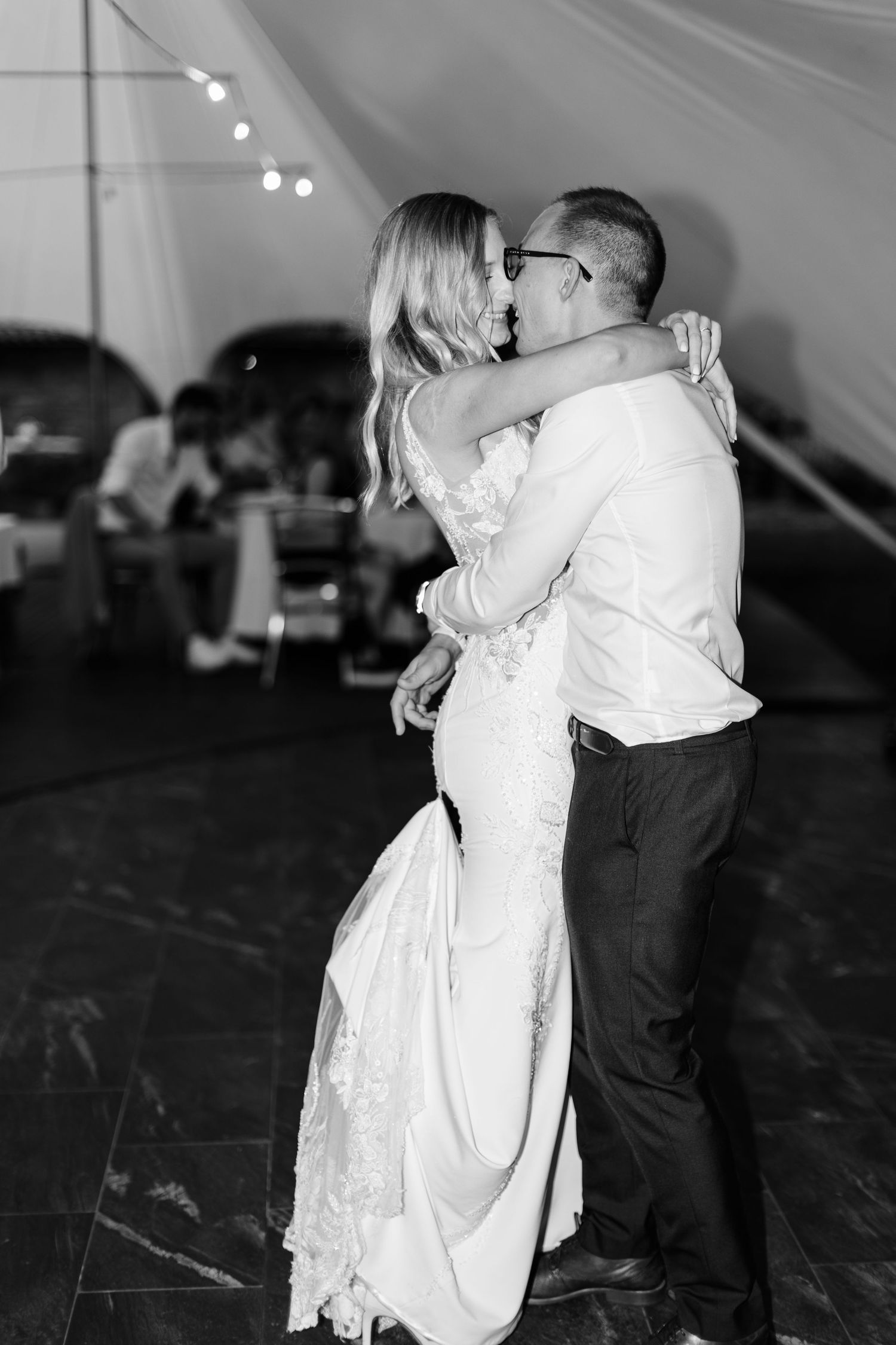 Black and white photo of bride and groom sharing first dance under string lights at elegant wedding reception.