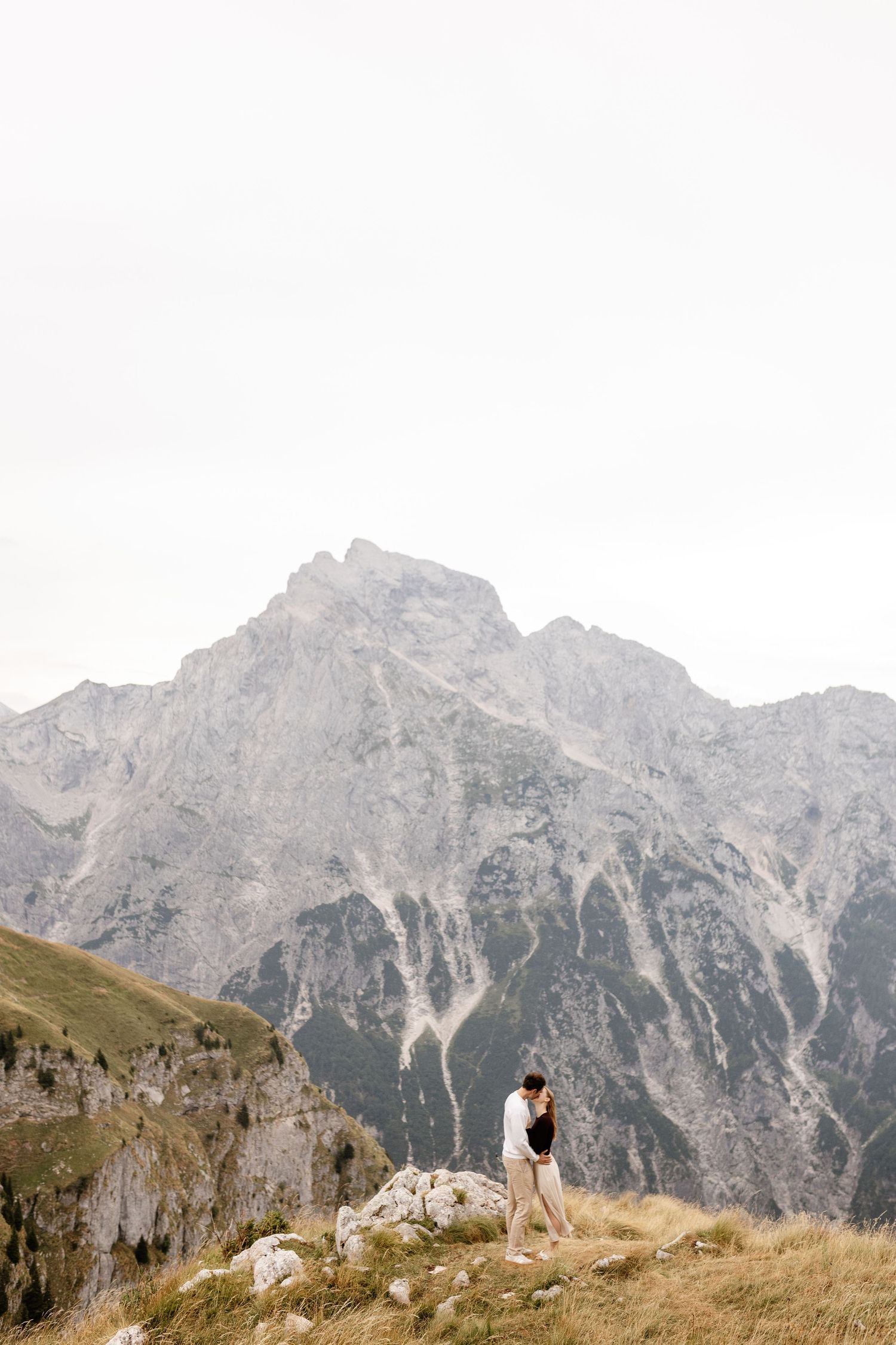 Couple embracing on a rocky mountain ridge with dramatic alpine peaks in the background.