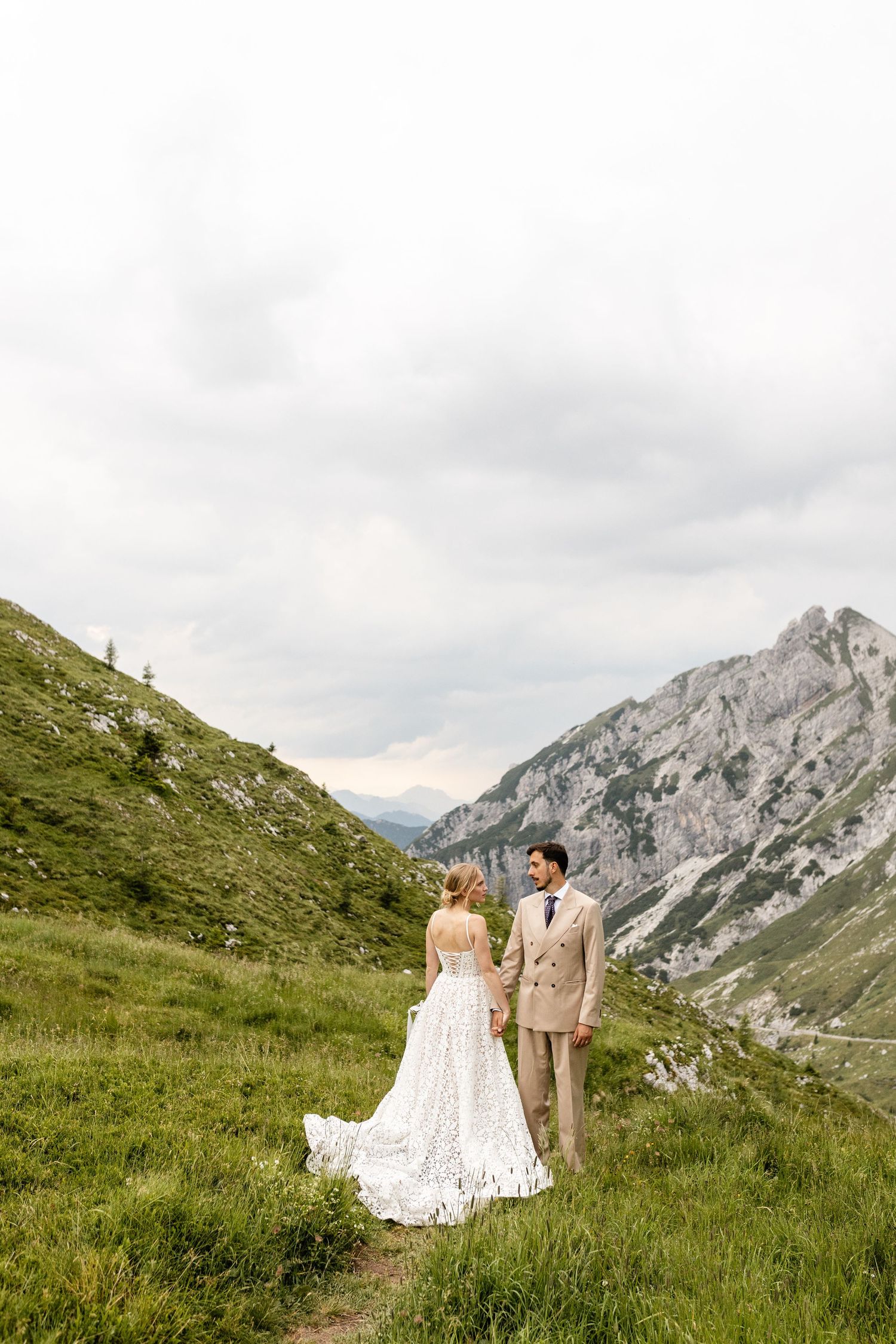 Bride and groom stand on a scenic mountain trail in the Alps, surrounded by lush green hills and dramatic rocky peaks.
