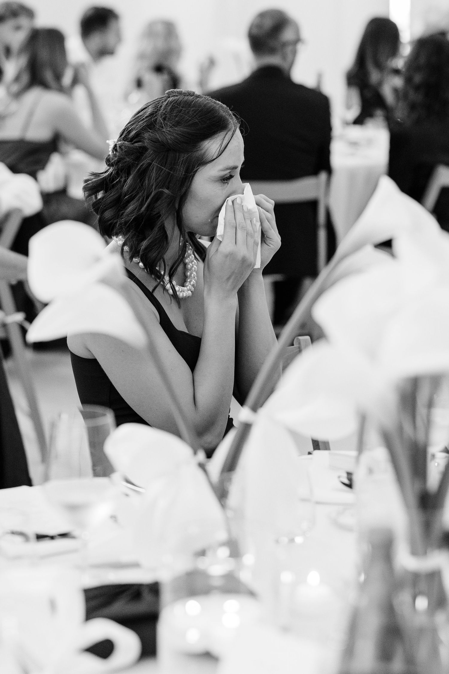 Emotional wedding guest in black dress dabs tears with tissue while seated at decorated reception table.