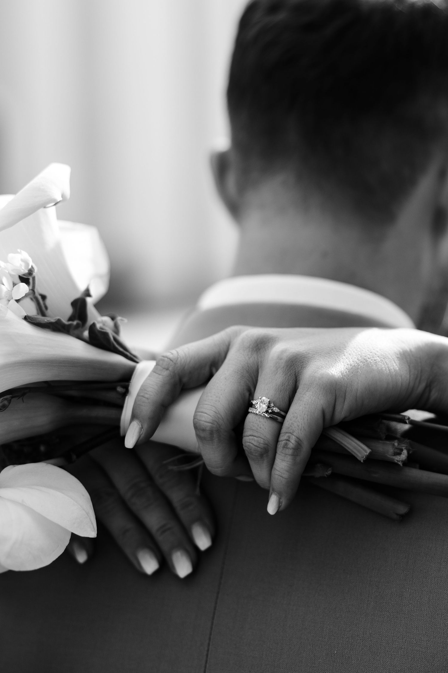 Black and white close-up of bride's hand with diamond ring resting on groom's shoulder.