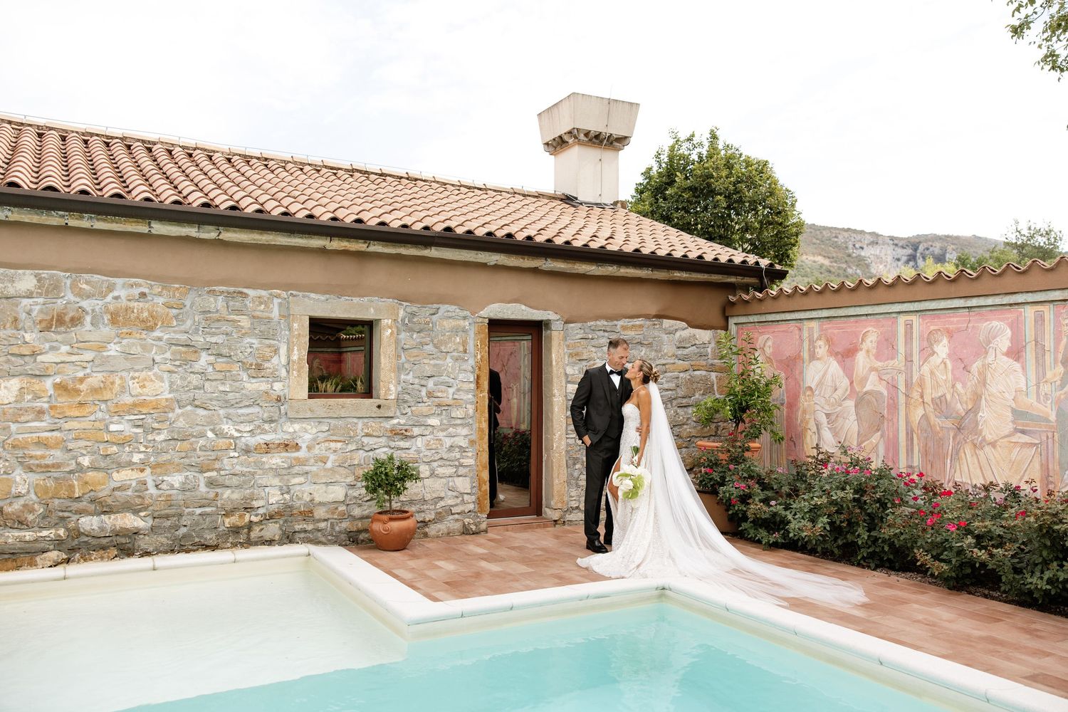 Bride and groom pose romantically by a pool at a stone villa with a colorful mural backdrop.