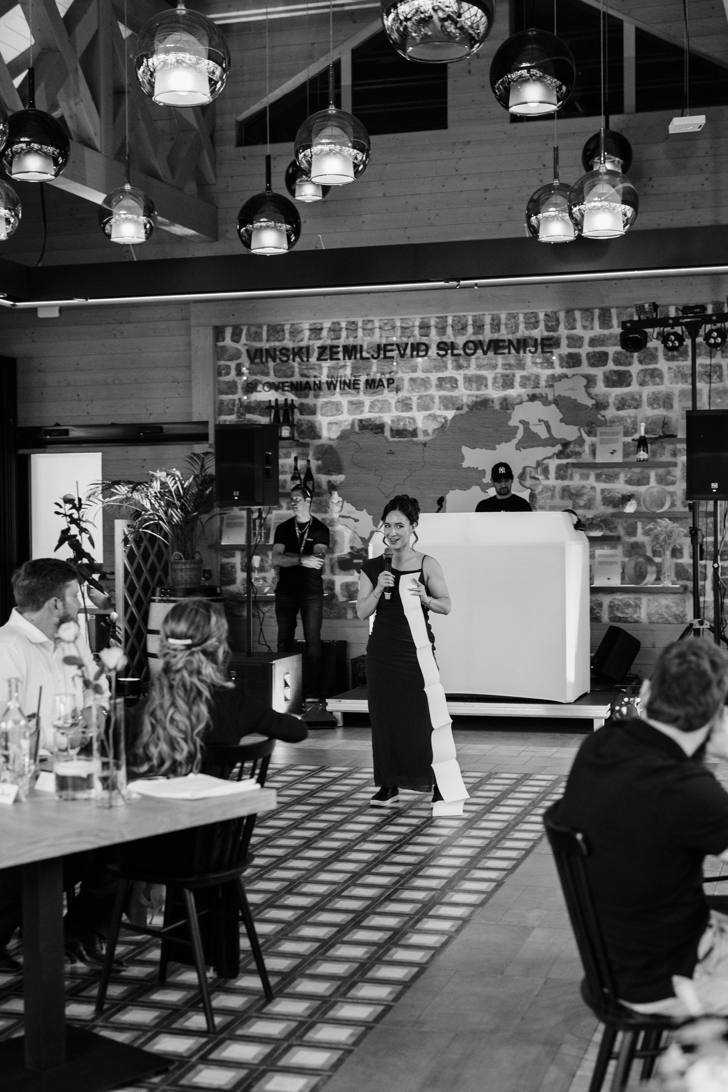 Wedding guest delivers a speech at a rustic venue with pendant lights and a Slovenian map mural backdrop.