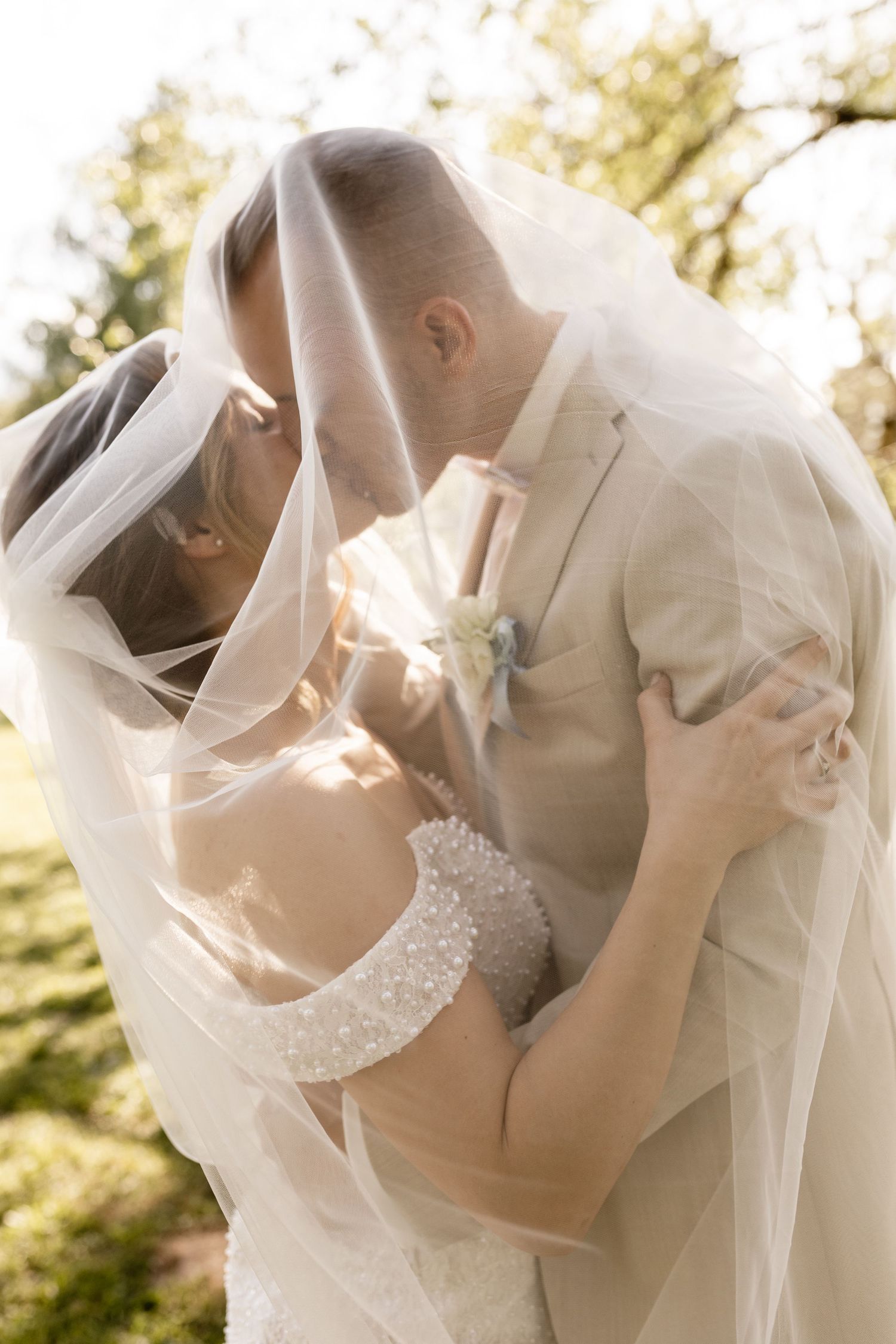 Bride and groom sharing a romantic kiss under a flowing wedding veil outdoors in soft natural light.