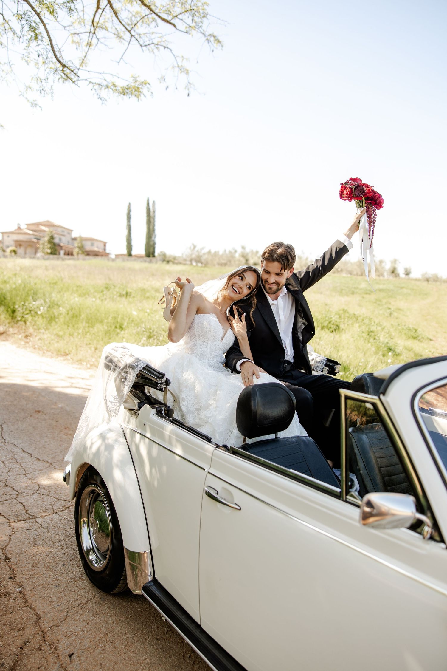 Joyful newlyweds celebrating in a white vintage convertible, bride in white gown, groom in suit holding red bouquet.
