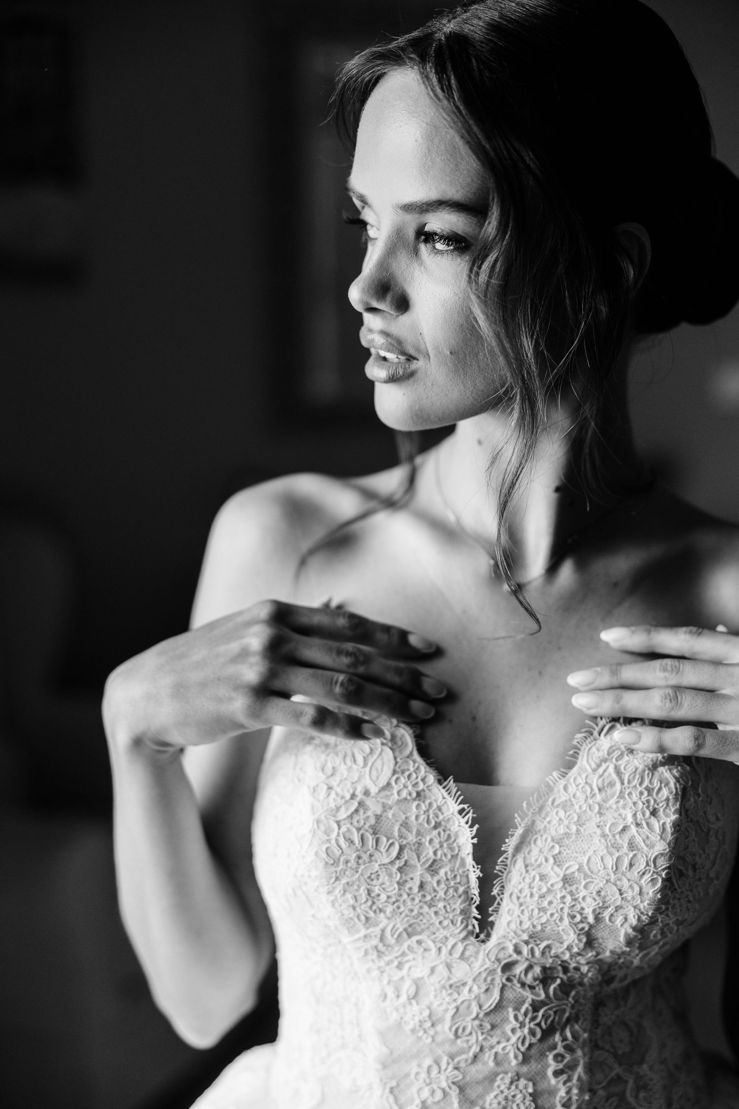 Black and white portrait of a bride in lace wedding dress adjusting her gown, gazing thoughtfully to the side.