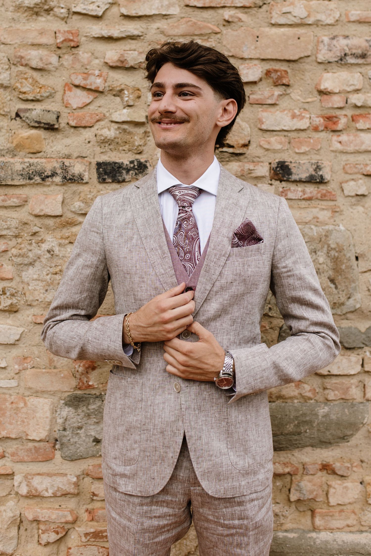 Smiling groom in beige linen suit with floral tie and pocket square stands against rustic brick wall.