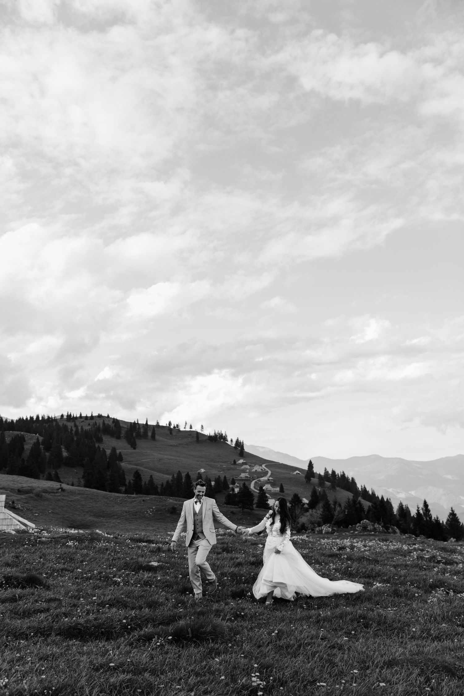 Black and white photo of a bride and groom holding hands on a scenic mountain meadow with evergreen trees and cloudy sky.