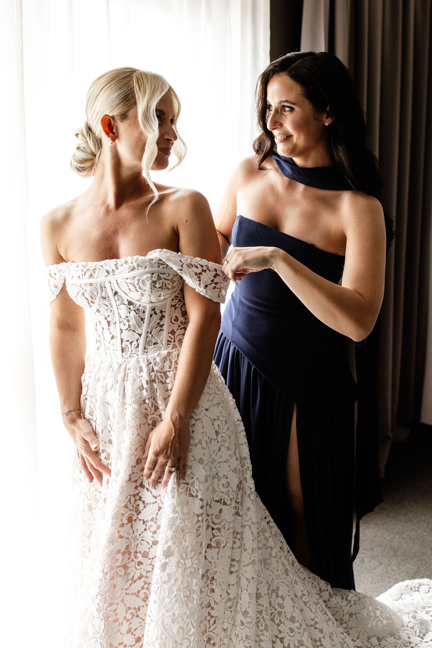 Bride in off-shoulder lace wedding gown being helped by bridesmaid in navy halter dress near window.