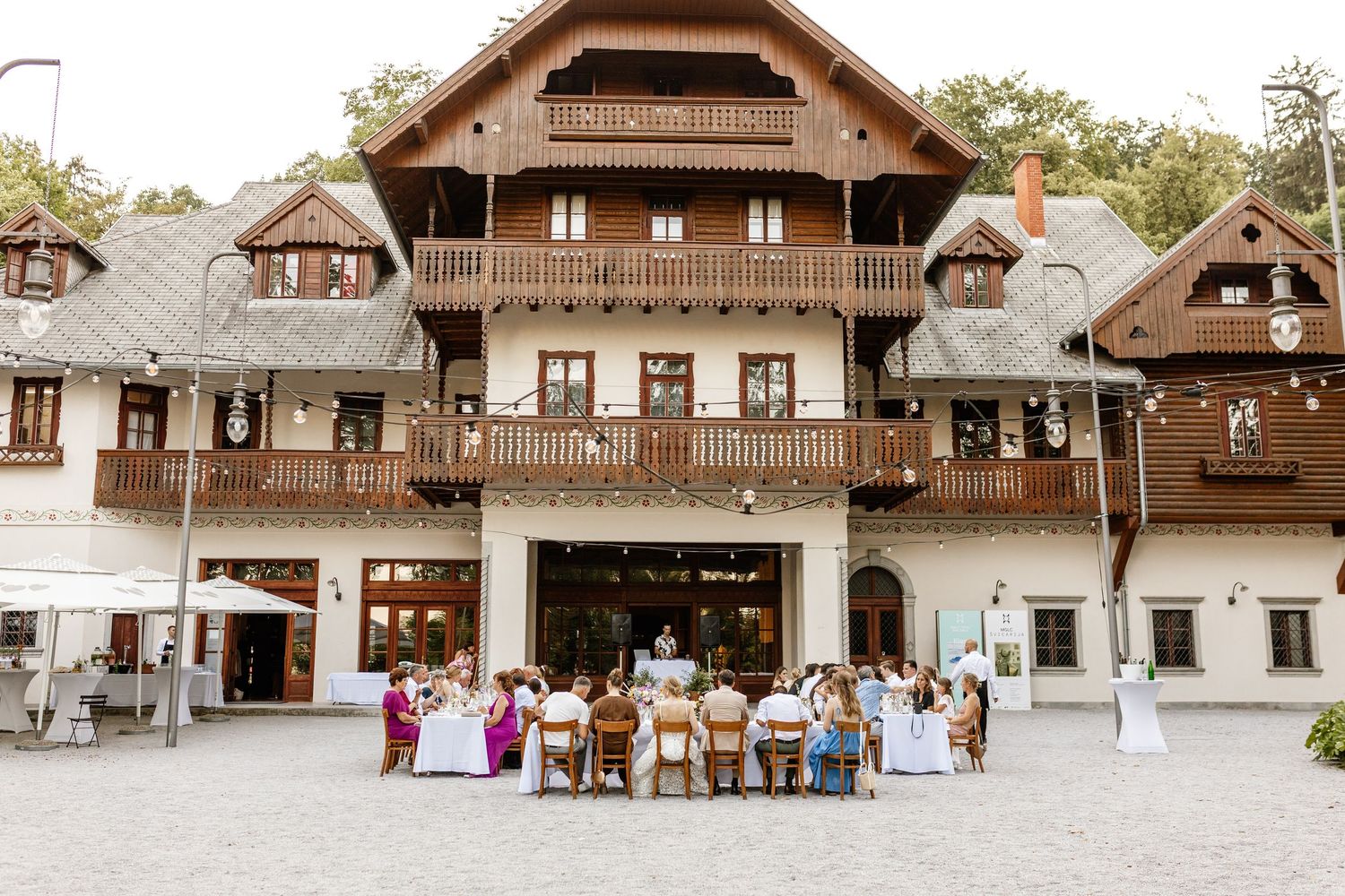 Elegant alpine-style wedding venue with guests seated at outdoor reception tables in front of ornate wooden building.