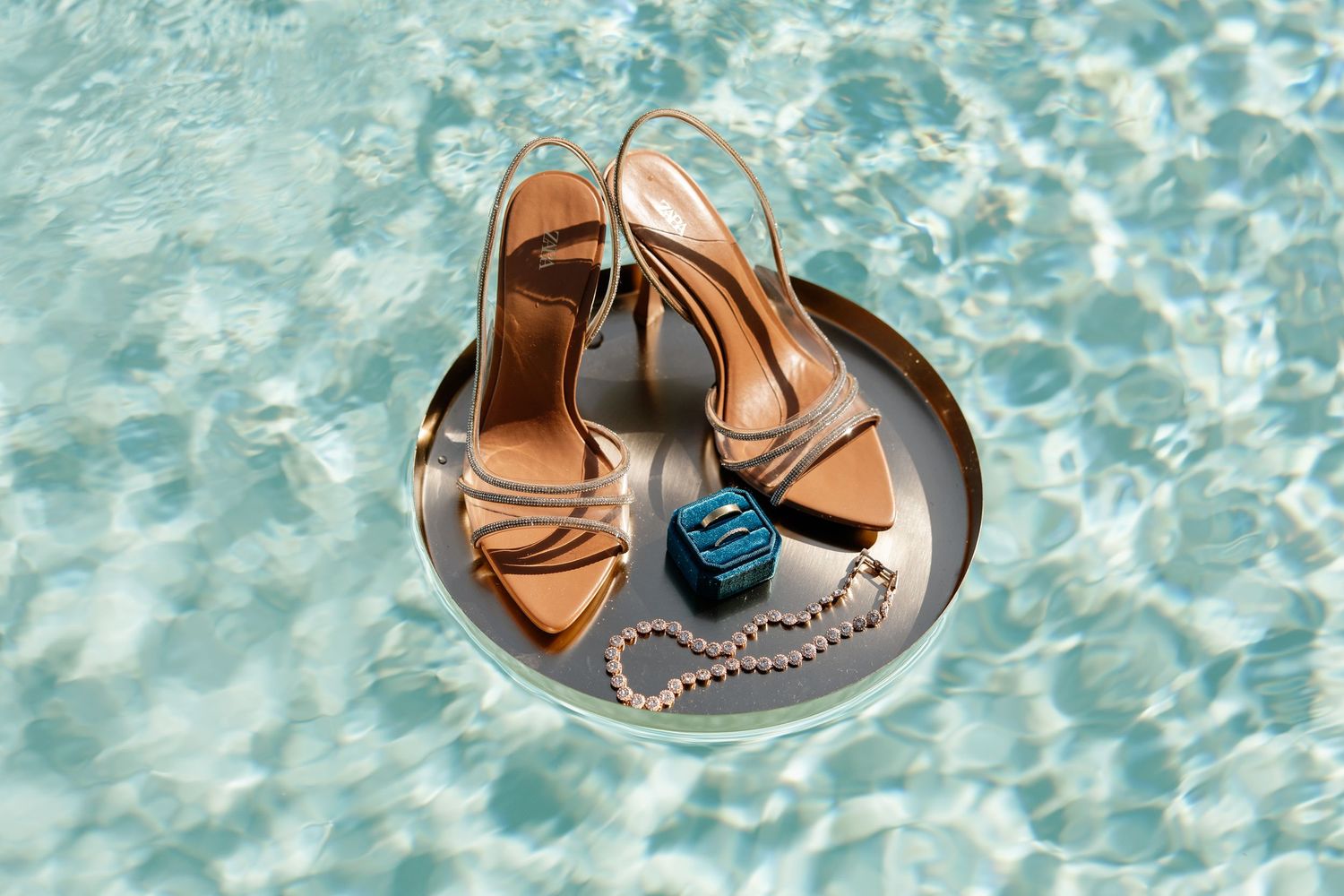 Gold strappy heeled sandals and blue jewelry on a silver tray floating in a turquoise pool.