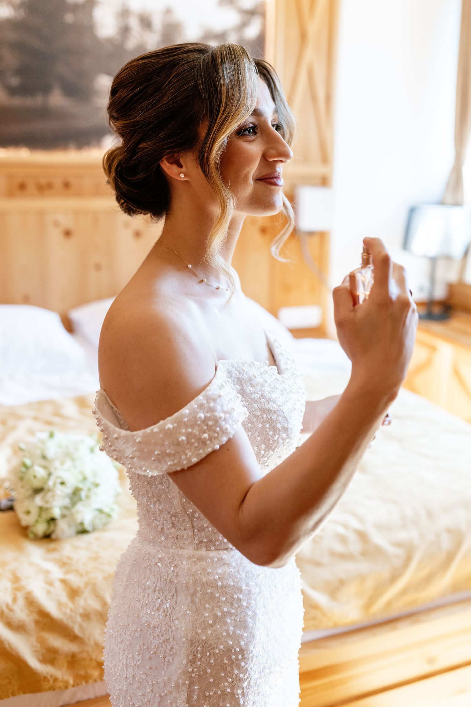 Elegant bride in a pearl-embellished off-shoulder wedding dress holding perfume in a warm wooden room.