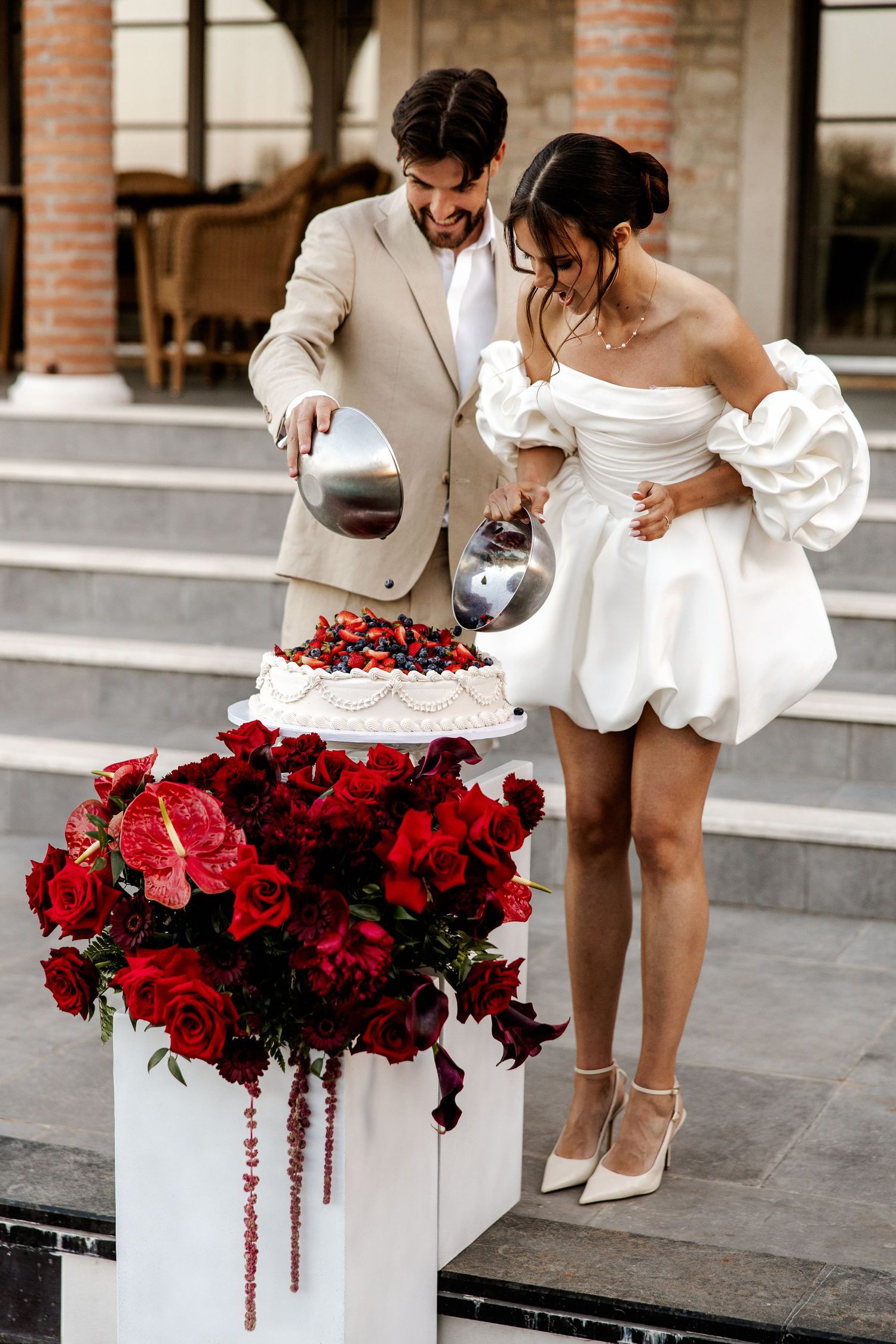 Smiling couple cutting a berry-topped wedding cake beside a lush red floral arrangement on a white pedestal.