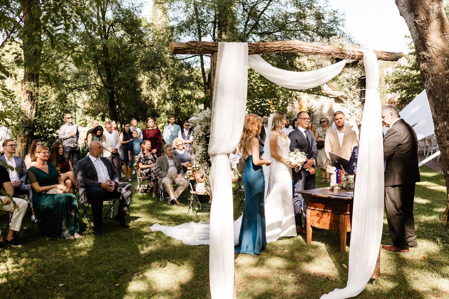 Outdoor wedding ceremony under rustic wooden arch with white draping, bride and groom before officiant, guests seated on lawn.