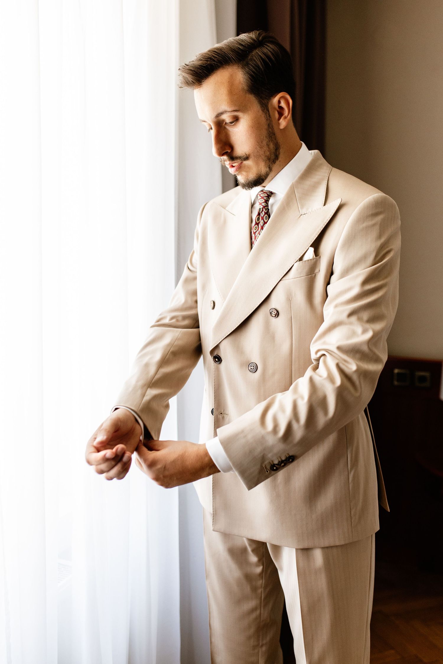 Elegant groom in a beige double-breasted suit adjusting his cufflinks by a bright window.