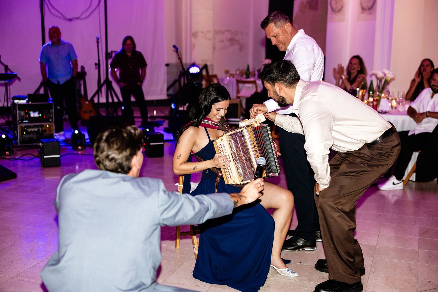 Woman playing accordion at wedding reception while guests dance around her in elegant ballroom.