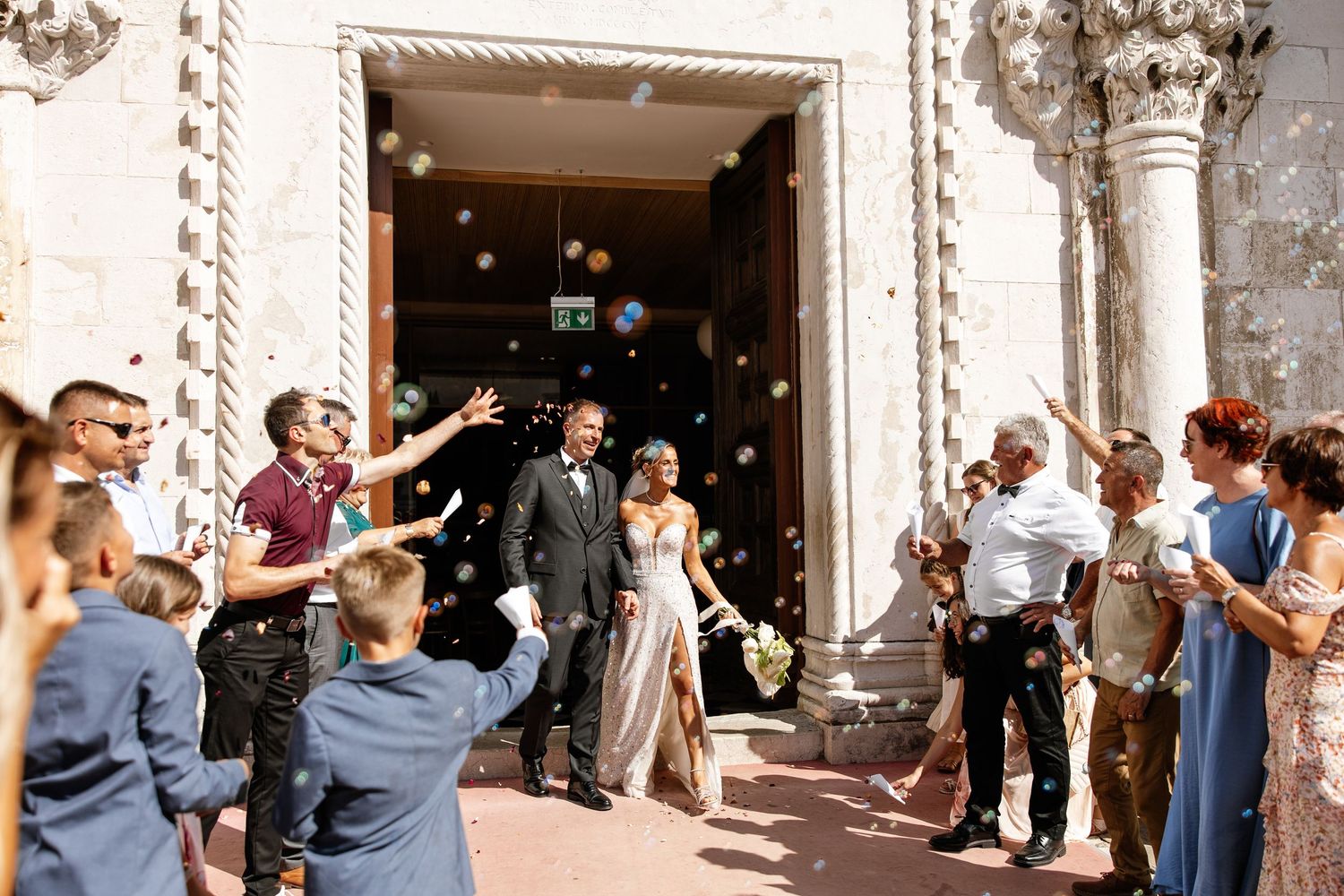 Bride and groom exit church as guests toss confetti during outdoor wedding ceremony.