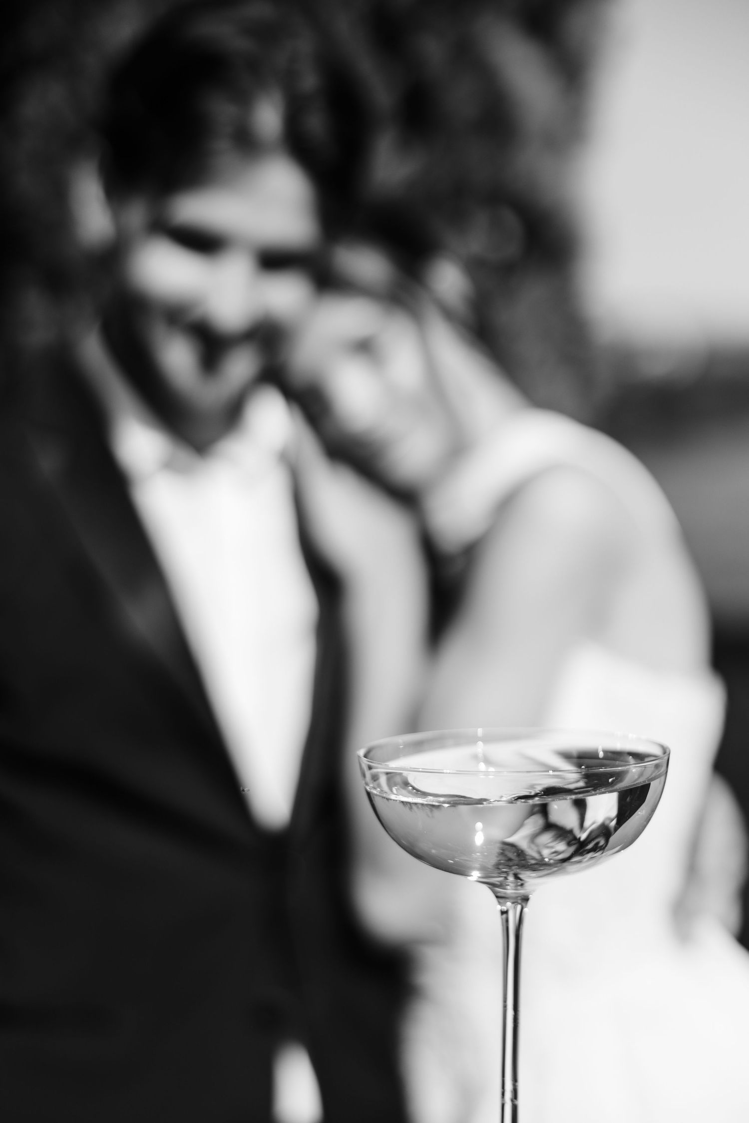 Black and white photo of newlyweds embracing behind a elegant champagne coupe glass in the foreground.