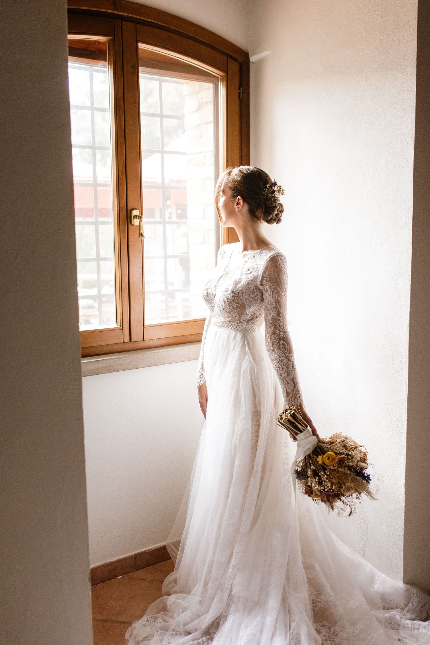 Elegant bride in long-sleeve lace wedding gown holds dried flower bouquet while gazing out a window.