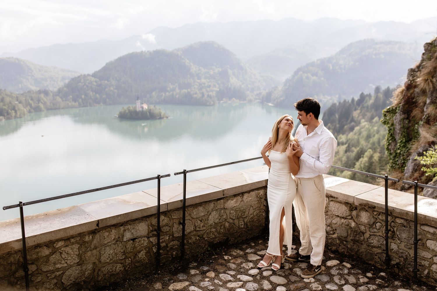 Couple embracing on stone terrace overlooking Lake Bled island and lush green mountains in Slovenia.
