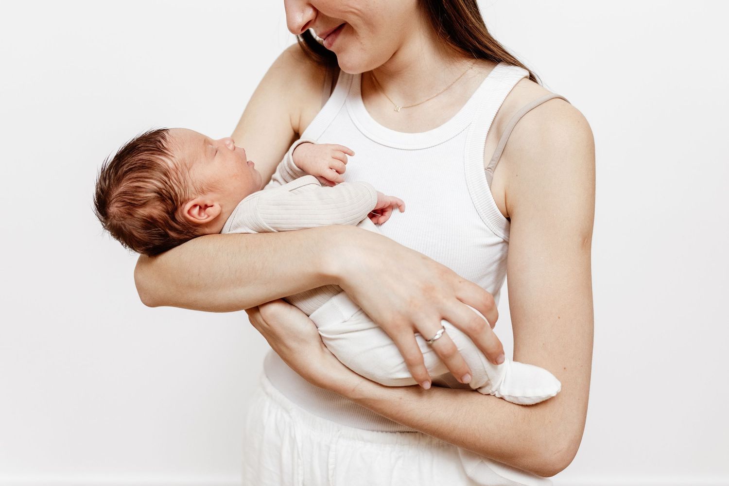 A smiling mother in a white tank top gently cradles her sleeping newborn baby against her chest.