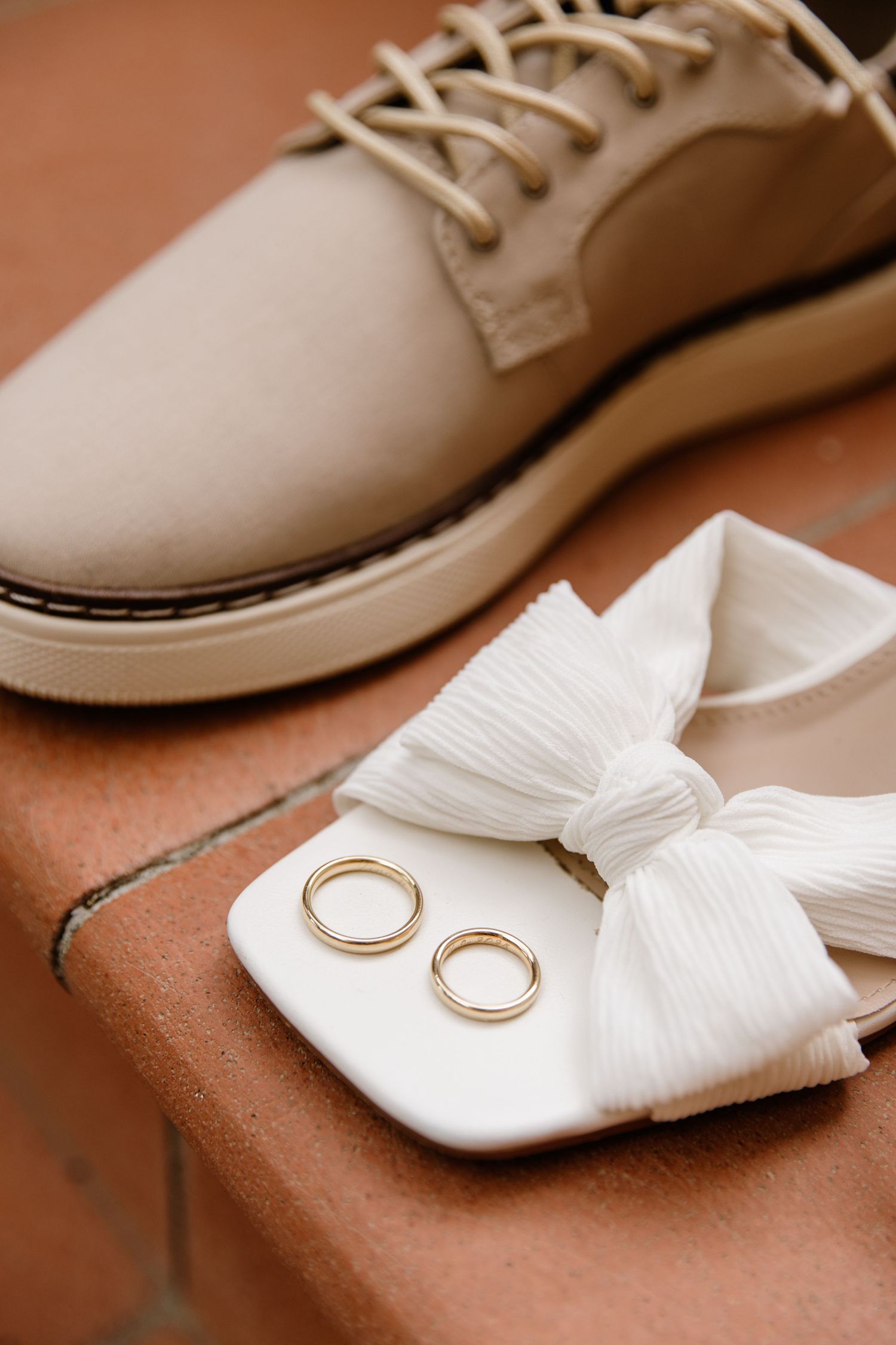 Two gold wedding bands rest on a white bow beside beige lace-up dress shoes on a brick surface.