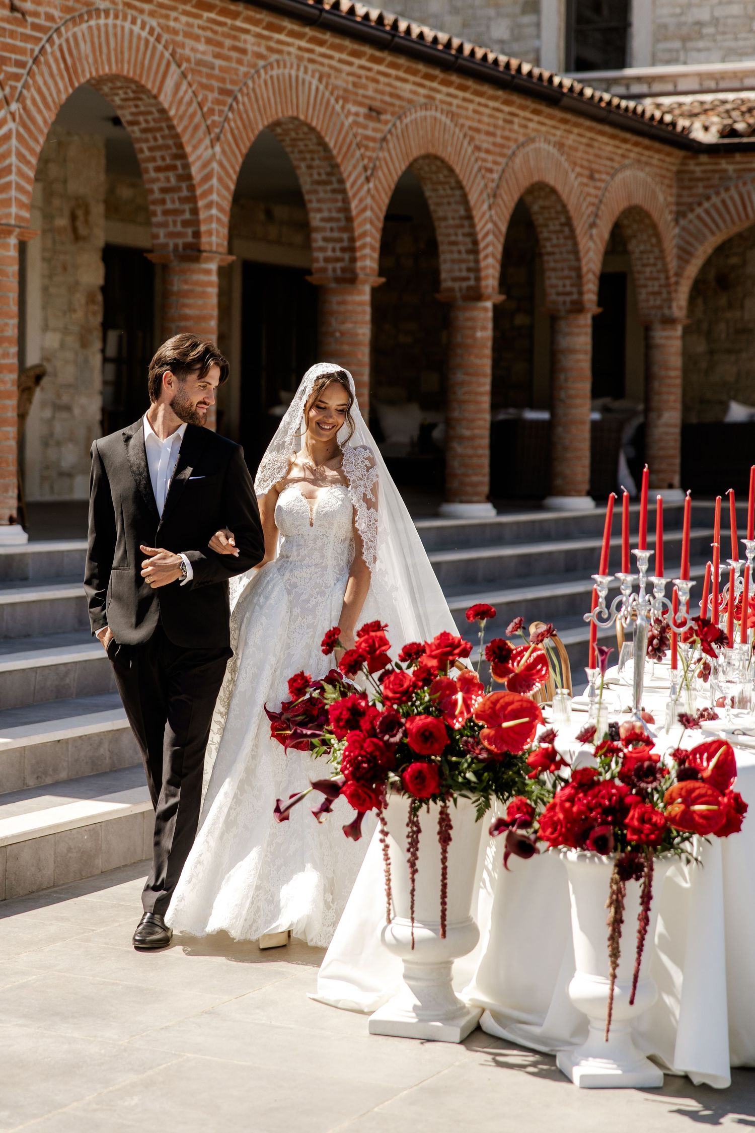 Elegant bride in white lace gown and groom in black suit walk past red floral arrangements at outdoor wedding venue.