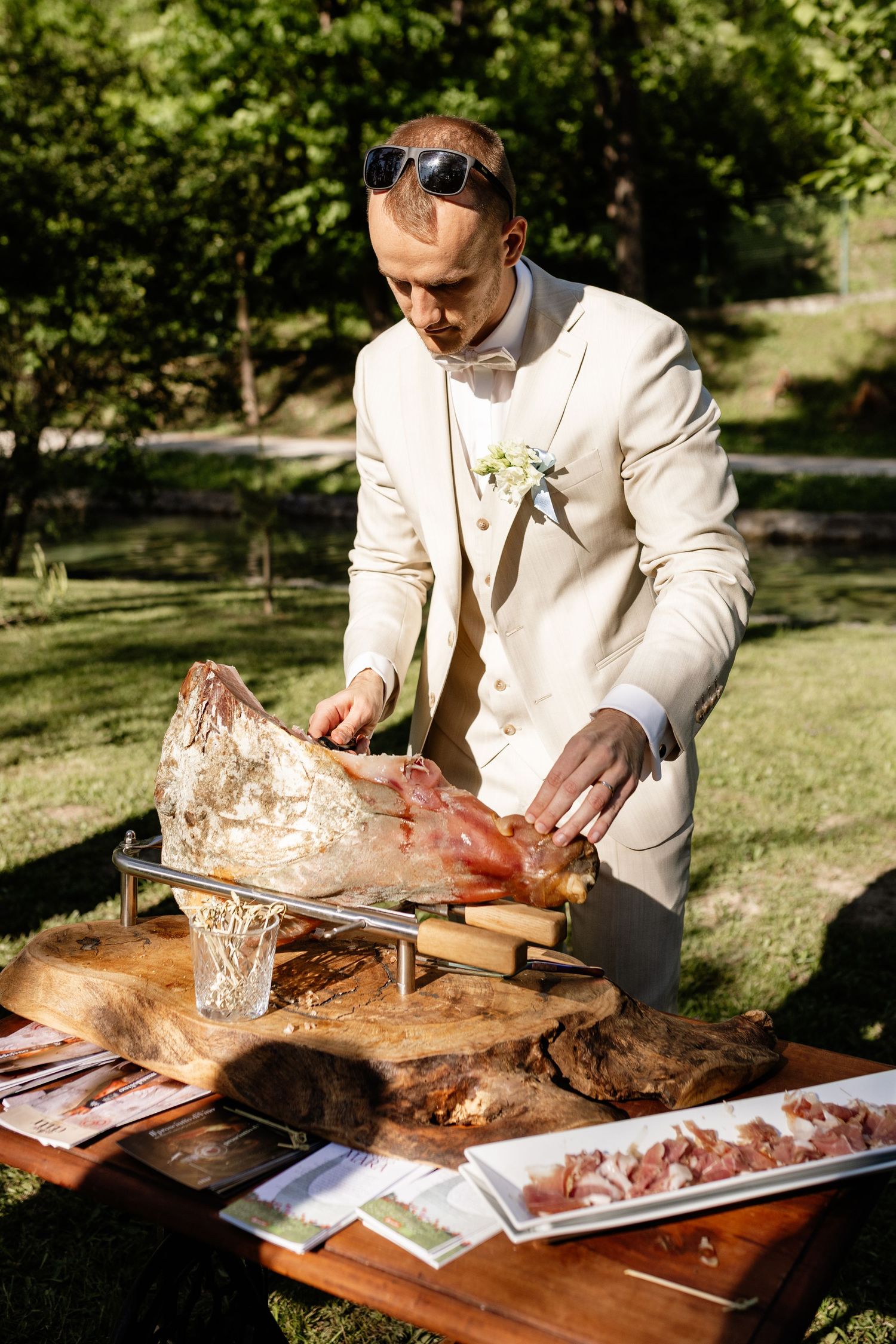 Elegant groom in white suit carving a whole leg of prosciutto on wooden board at outdoor wedding reception.
