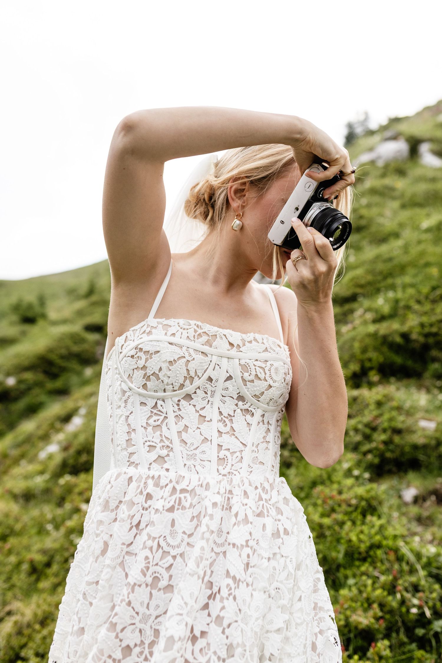 Blonde woman in white lace dress photographing nature with a film camera on a green hillside.