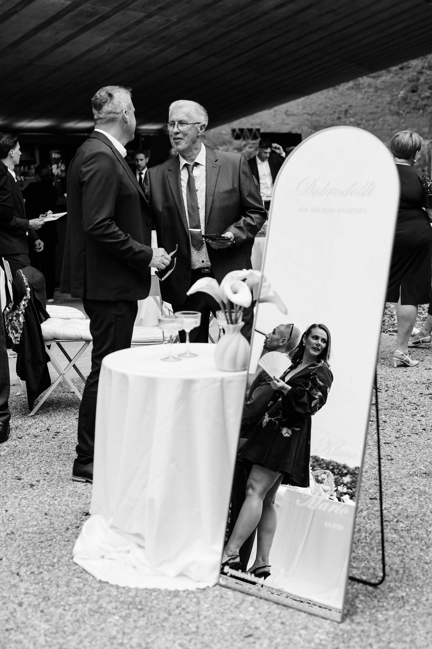Black and white photo of wedding guests socializing outdoors near a mirror and white cocktail table.