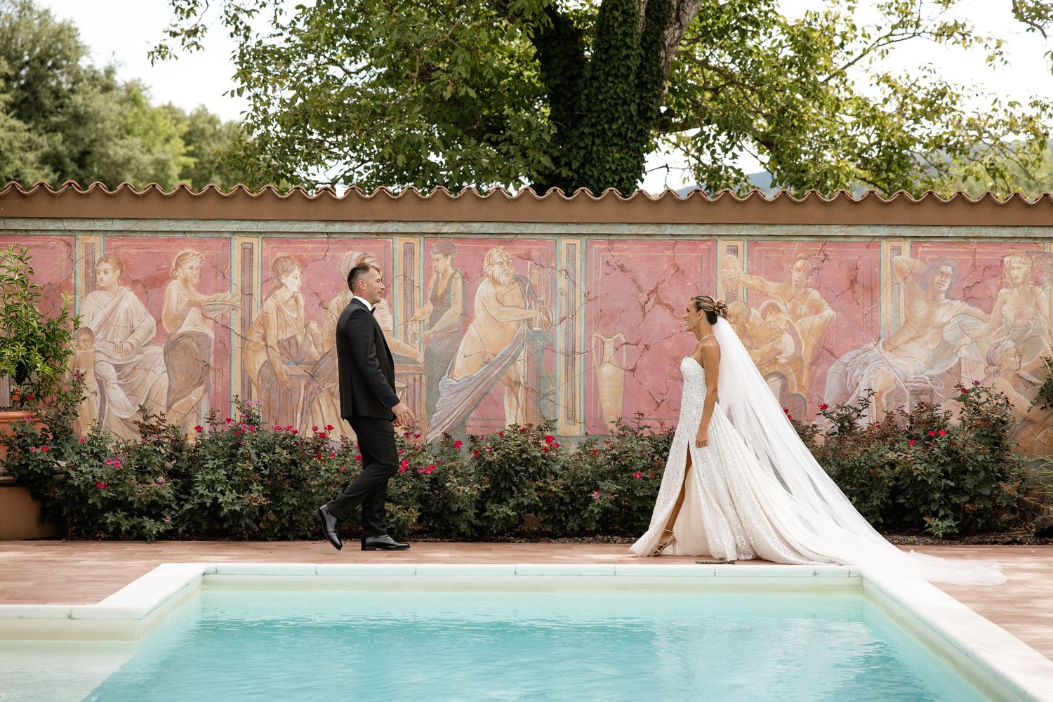 Bride and groom share first look moment beside pool with colorful Roman fresco mural backdrop outdoors.