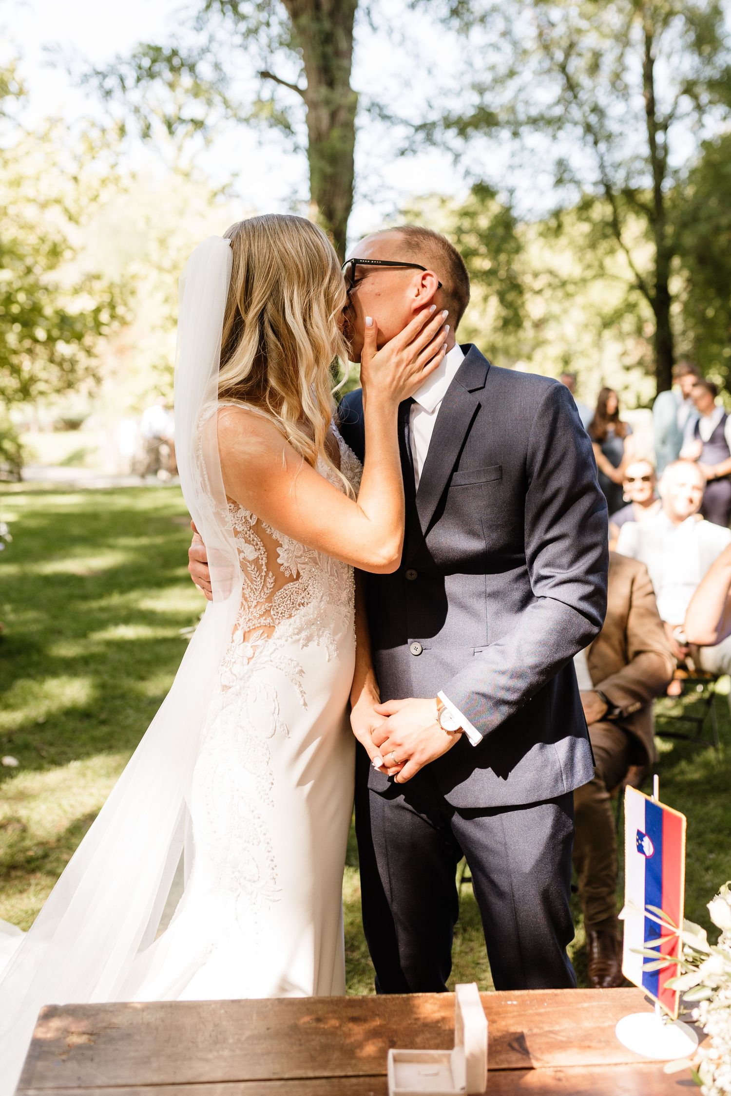 Bride and groom share their first kiss at an outdoor wedding ceremony surrounded by lush green trees.