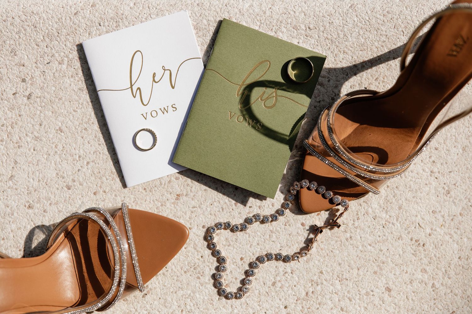 Bridal flat lay featuring tan heeled sandals, green vow book, wedding rings, and pearl necklace on neutral carpet.